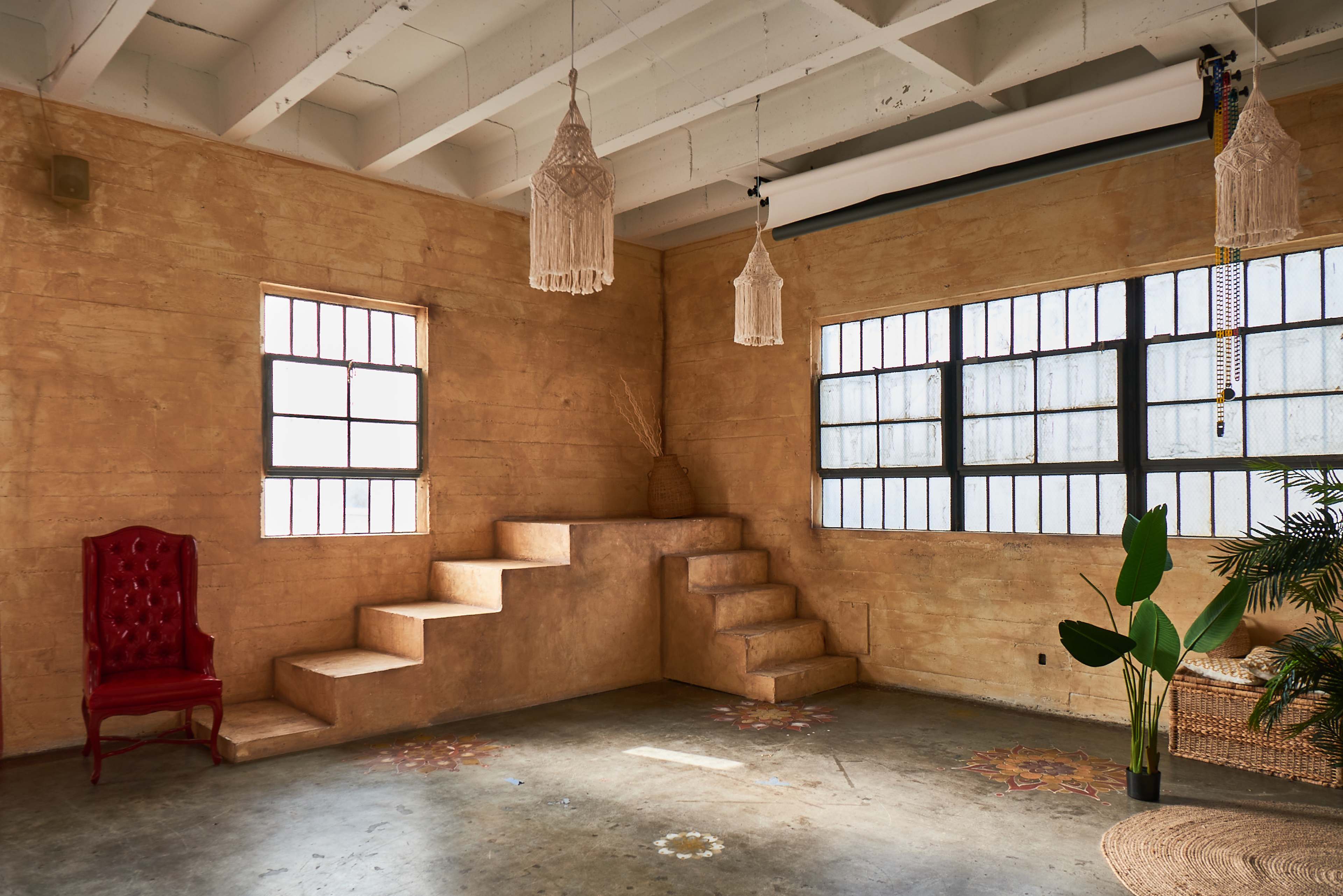 A well-lit, empty room with a textured yellow wall, two large windows, decorative hanging lights, a red chair, and a small staircase leading to an elevated platform.