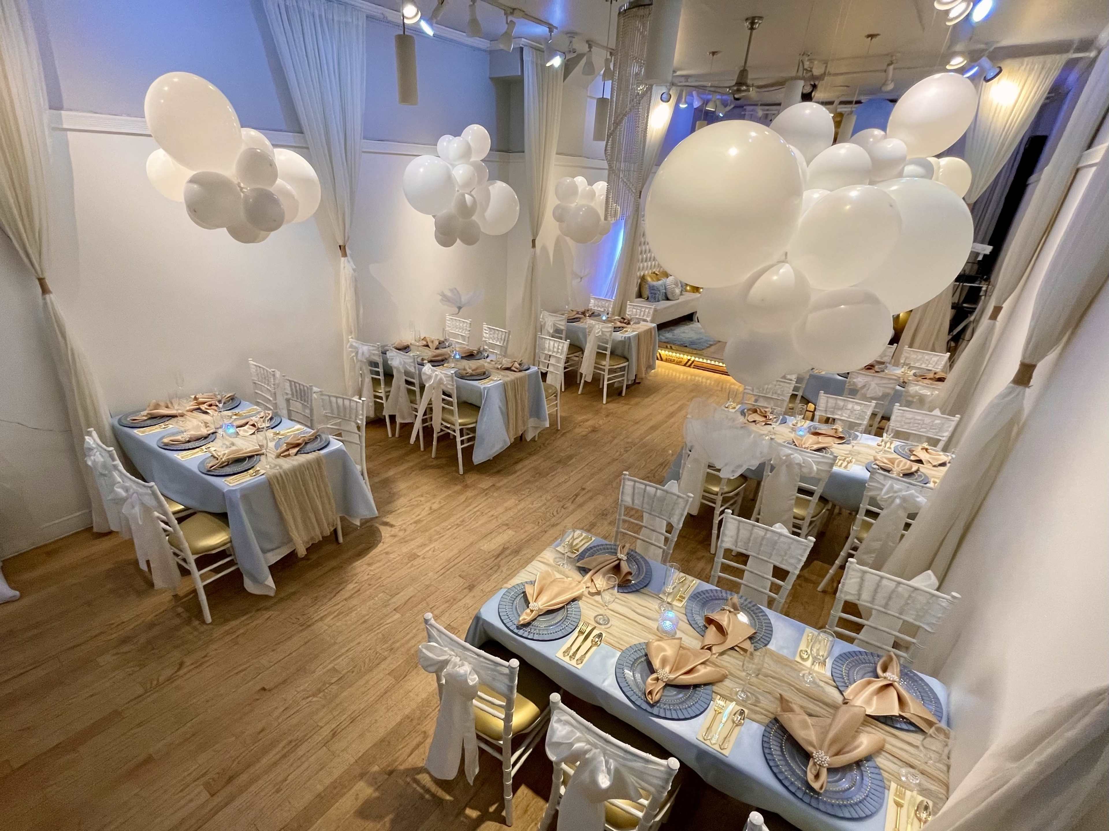 A banquet hall is set up with elegantly decorated tables that feature blue tablecloths, white chairs, and clusters of white balloons overhead.