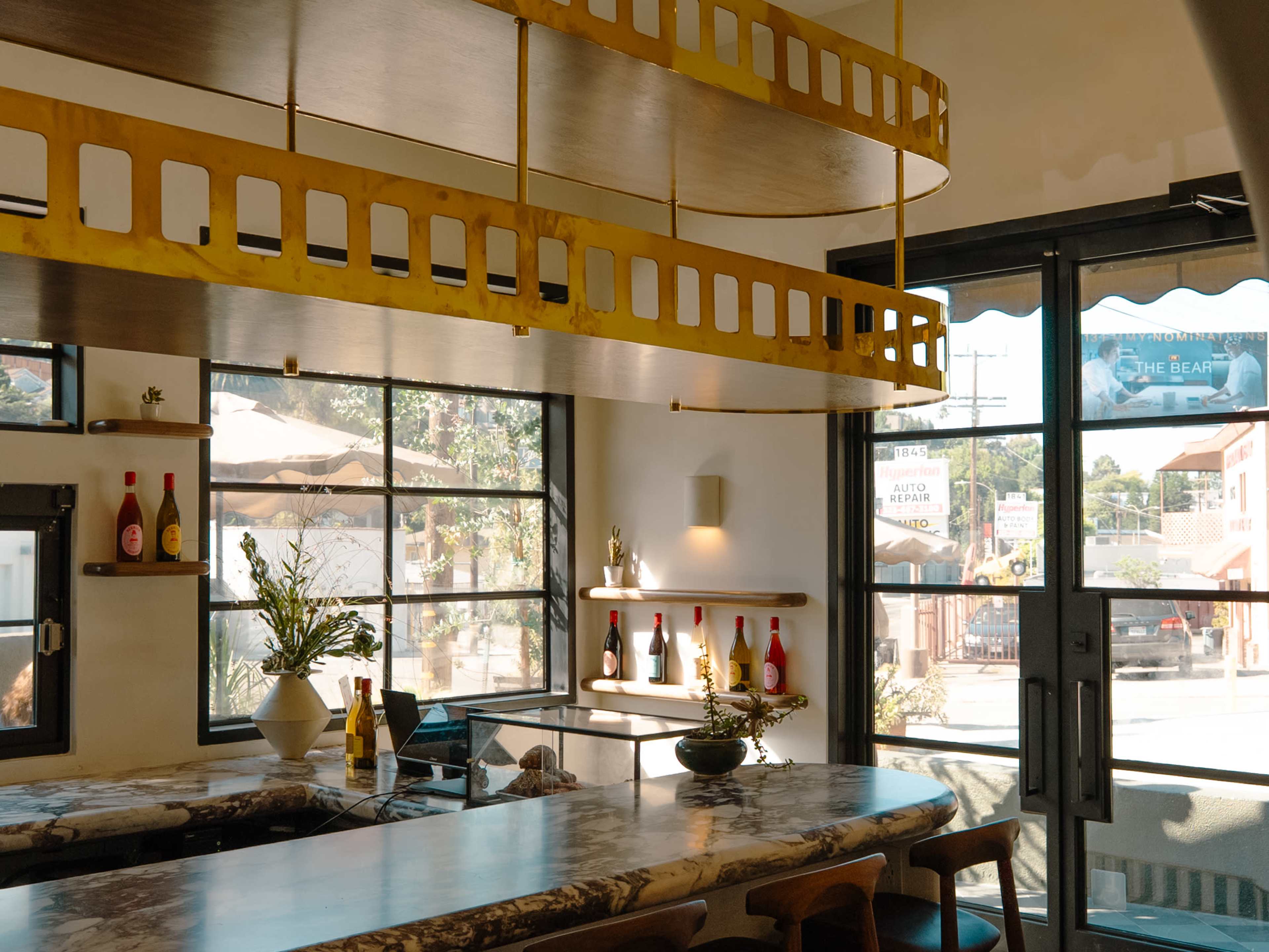 The image shows a modern interior of a café with large windows, a long marble bar, and decorative shelving displaying bottles and plants.