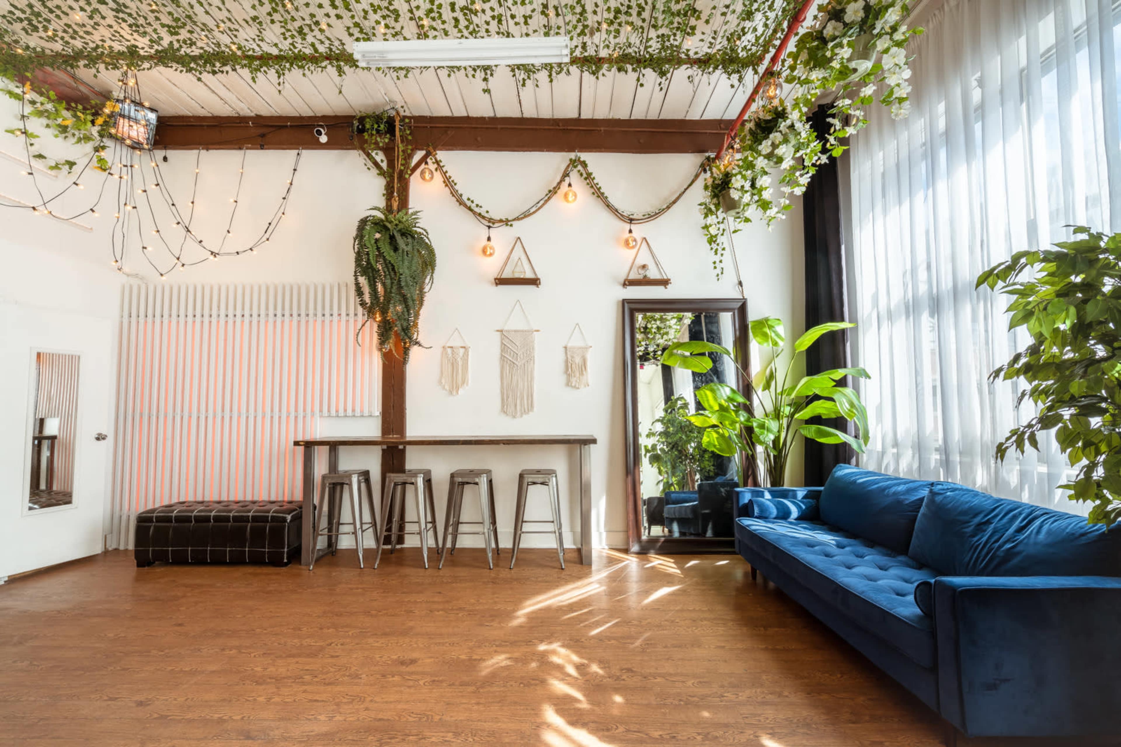 A bright interior space with a greenery-adorned ceiling, a wooden pillar, a long bar with stools, a large mirror, and a blue couch against the wall.