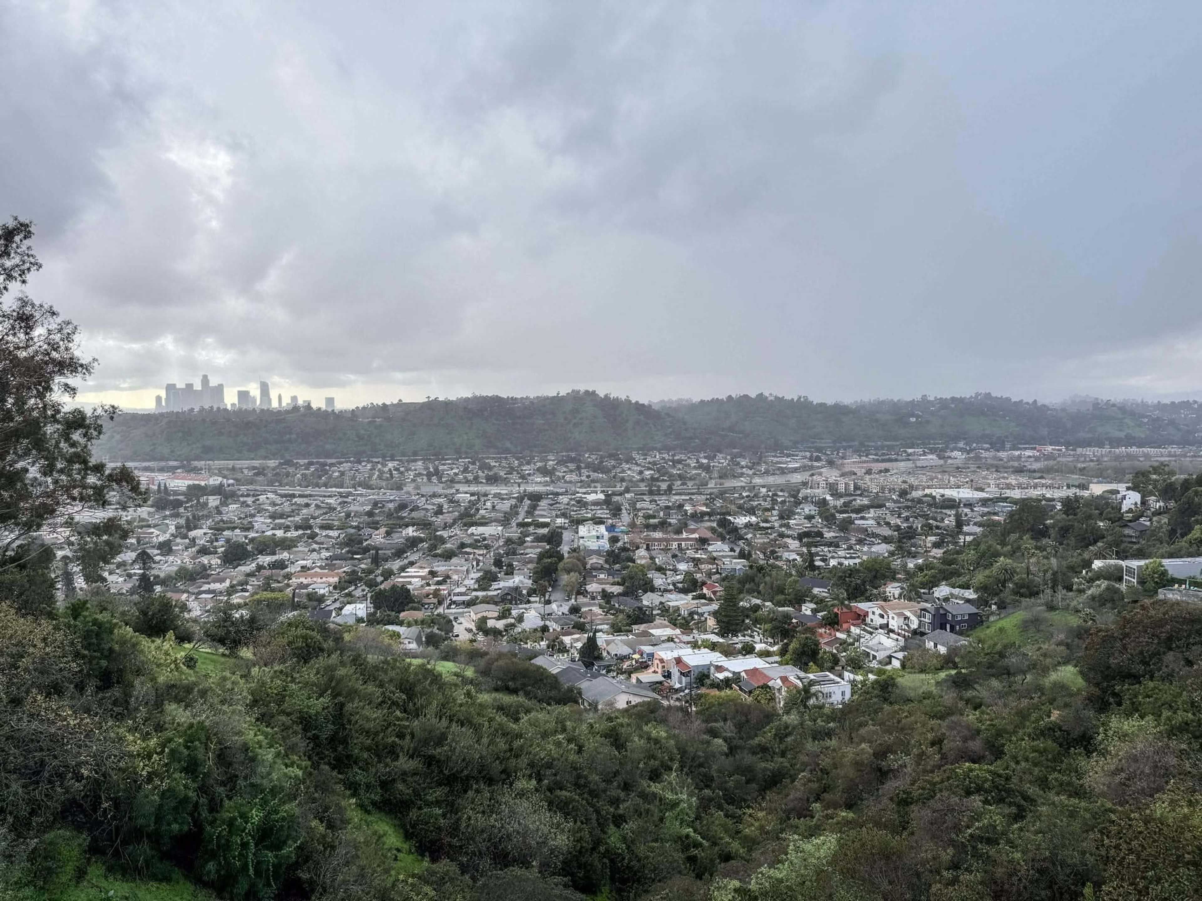 The image shows an aerial view of a densely populated neighborhood with a skyline in the distance, set against a backdrop of cloudy skies and rolling hills.