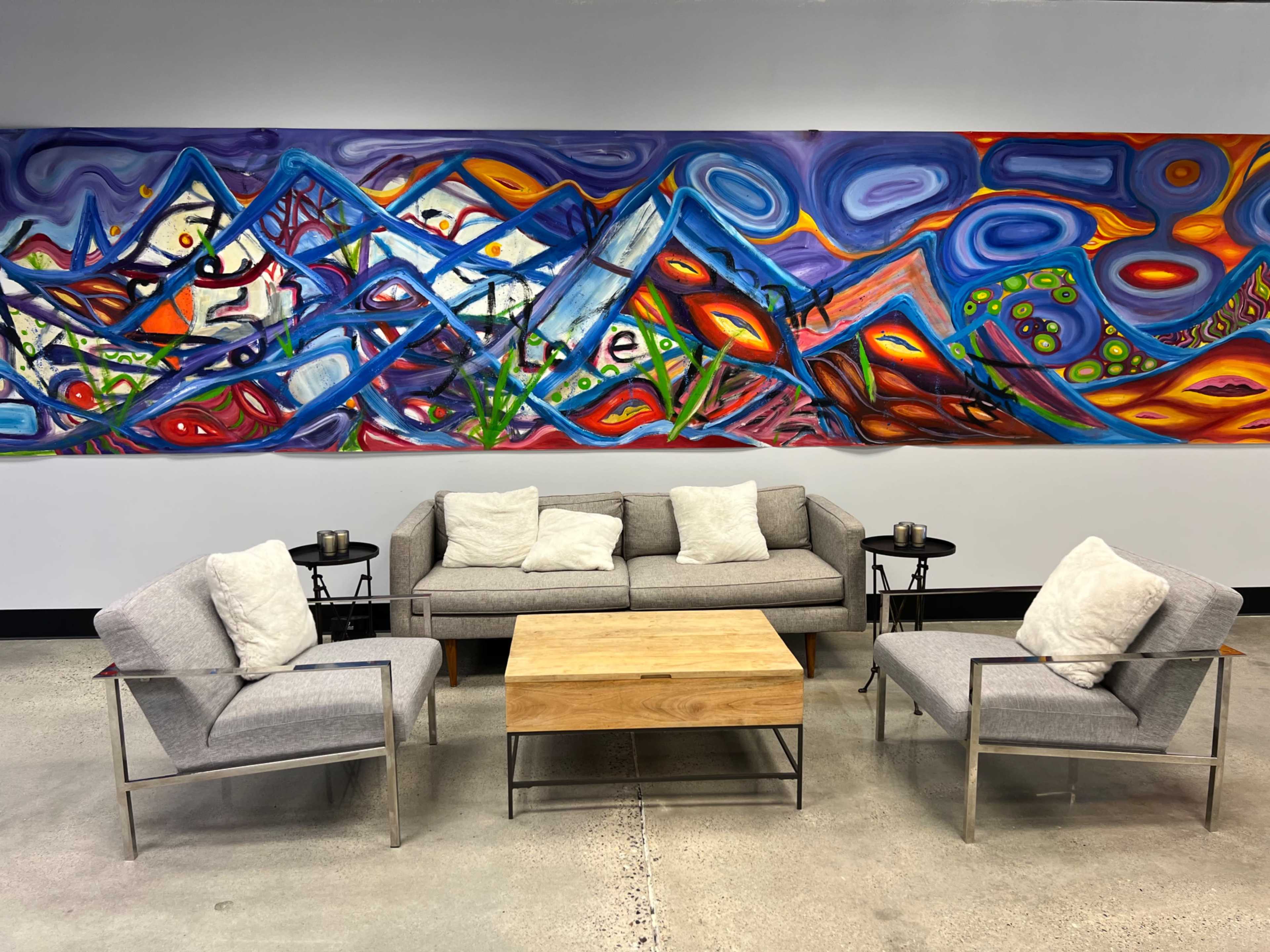 A gray fabric sofa and two armchairs are arranged around a wooden coffee table in front of a colorful abstract mural depicting mountains and patterns.