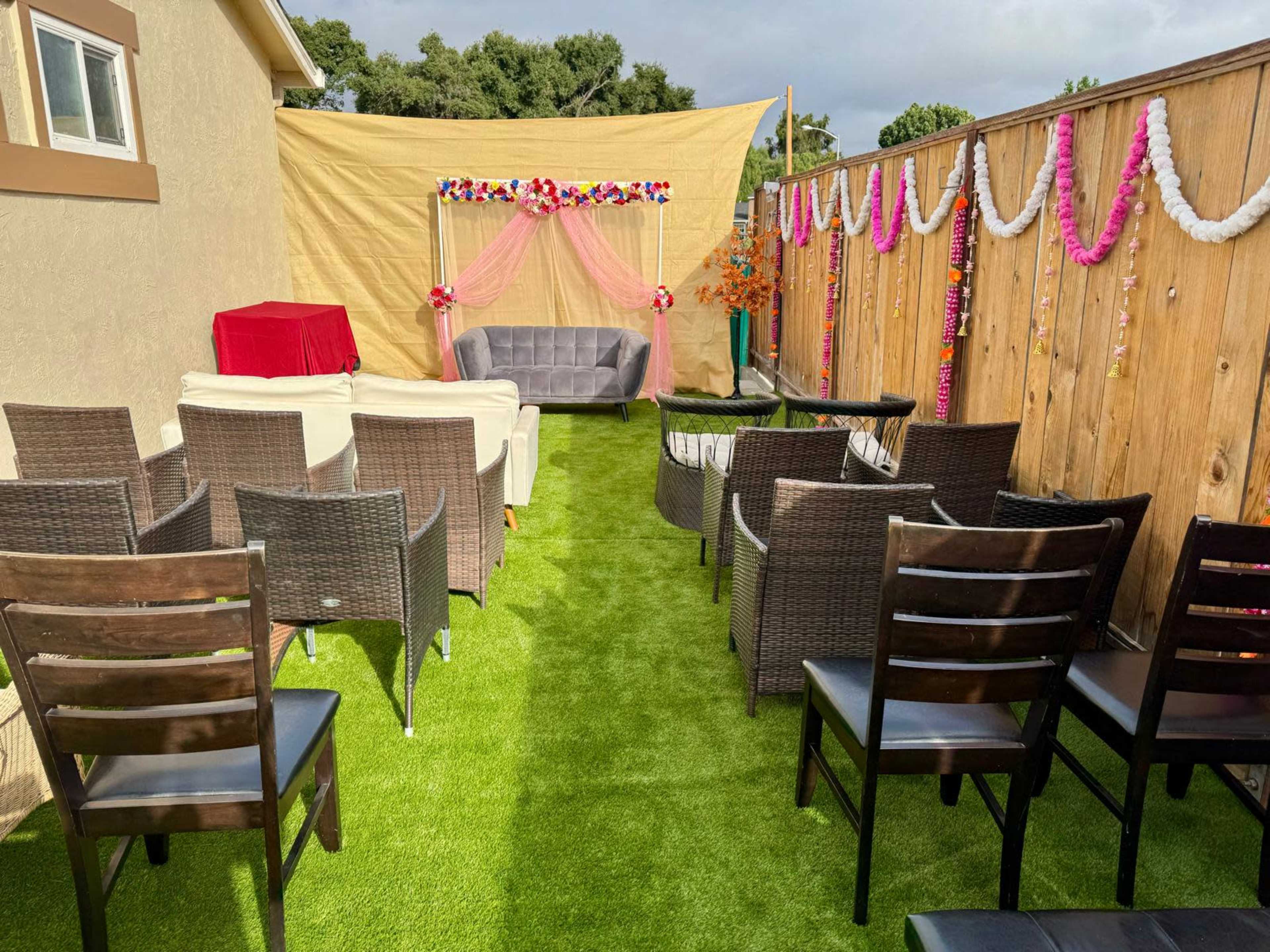 A well-decorated outdoor seating area with chairs organized in rows facing a couch and backdrop, surrounded by wooden fences and artificial grass.