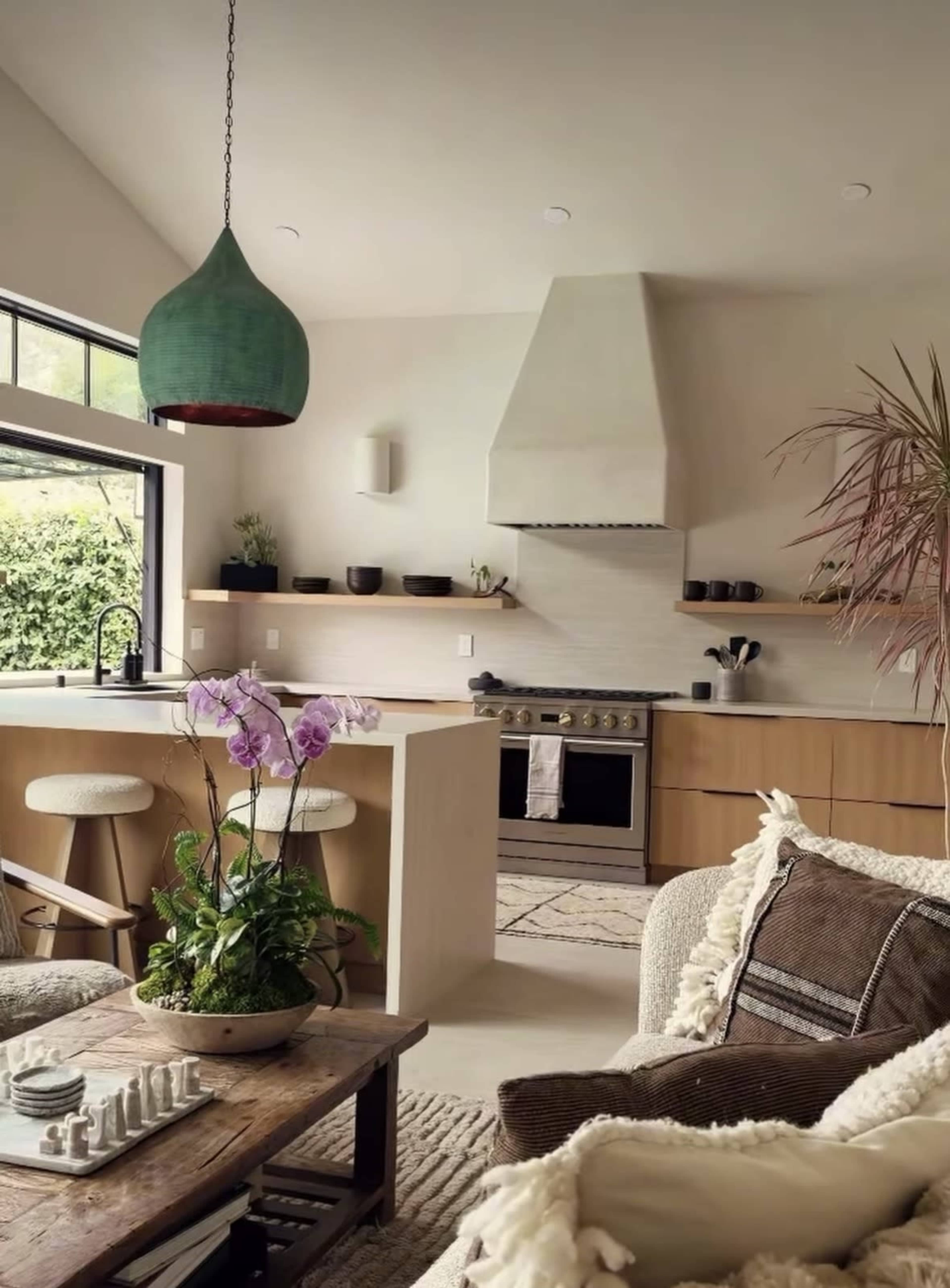 The image features a modern kitchen and living area with a wooden island, a stove, and a large window allowing natural light to illuminate the space.