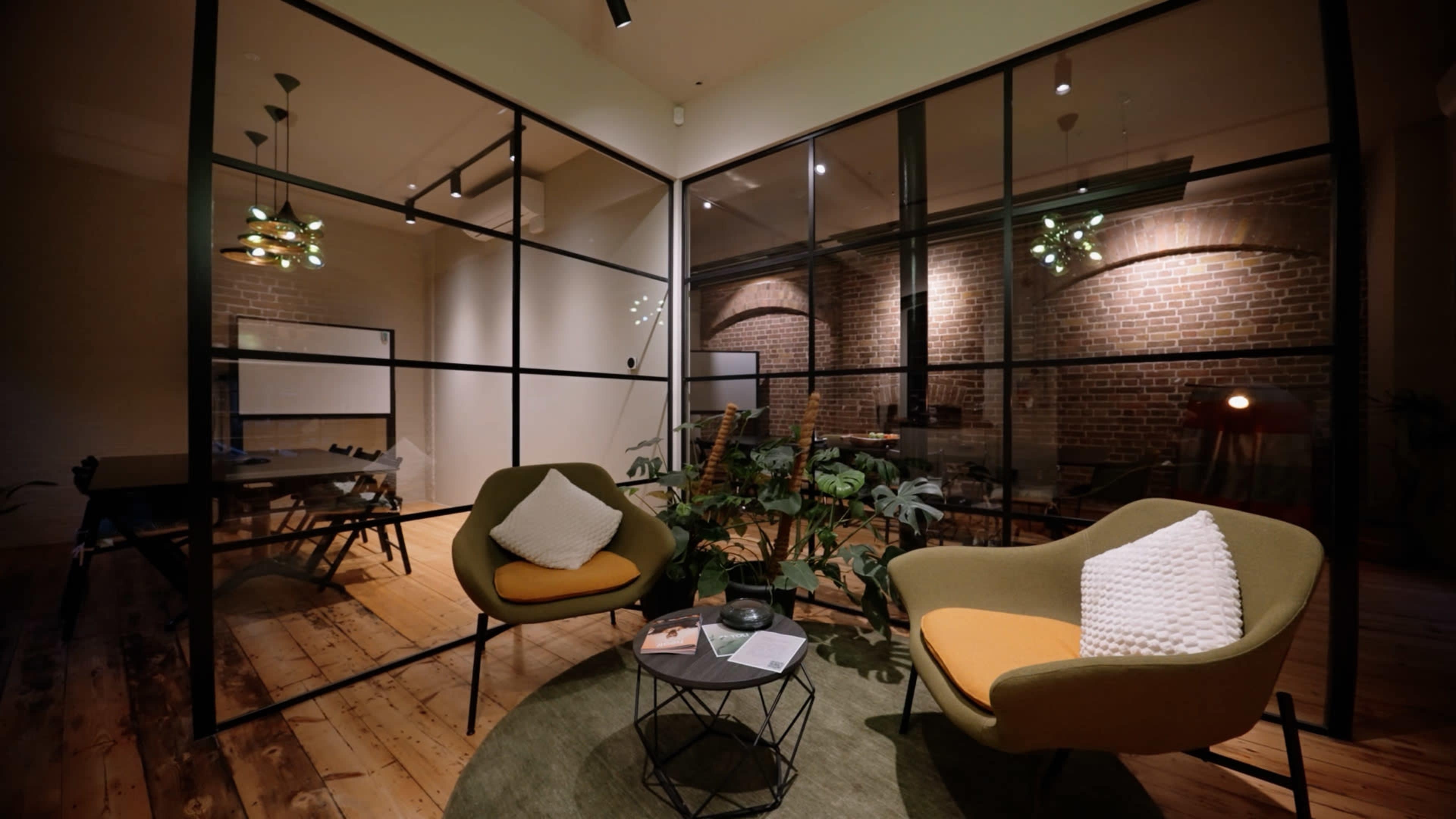 The image depicts a modern office space featuring two green chairs with orange accents, a small round table, and exposed brick walls in a glass-walled room.