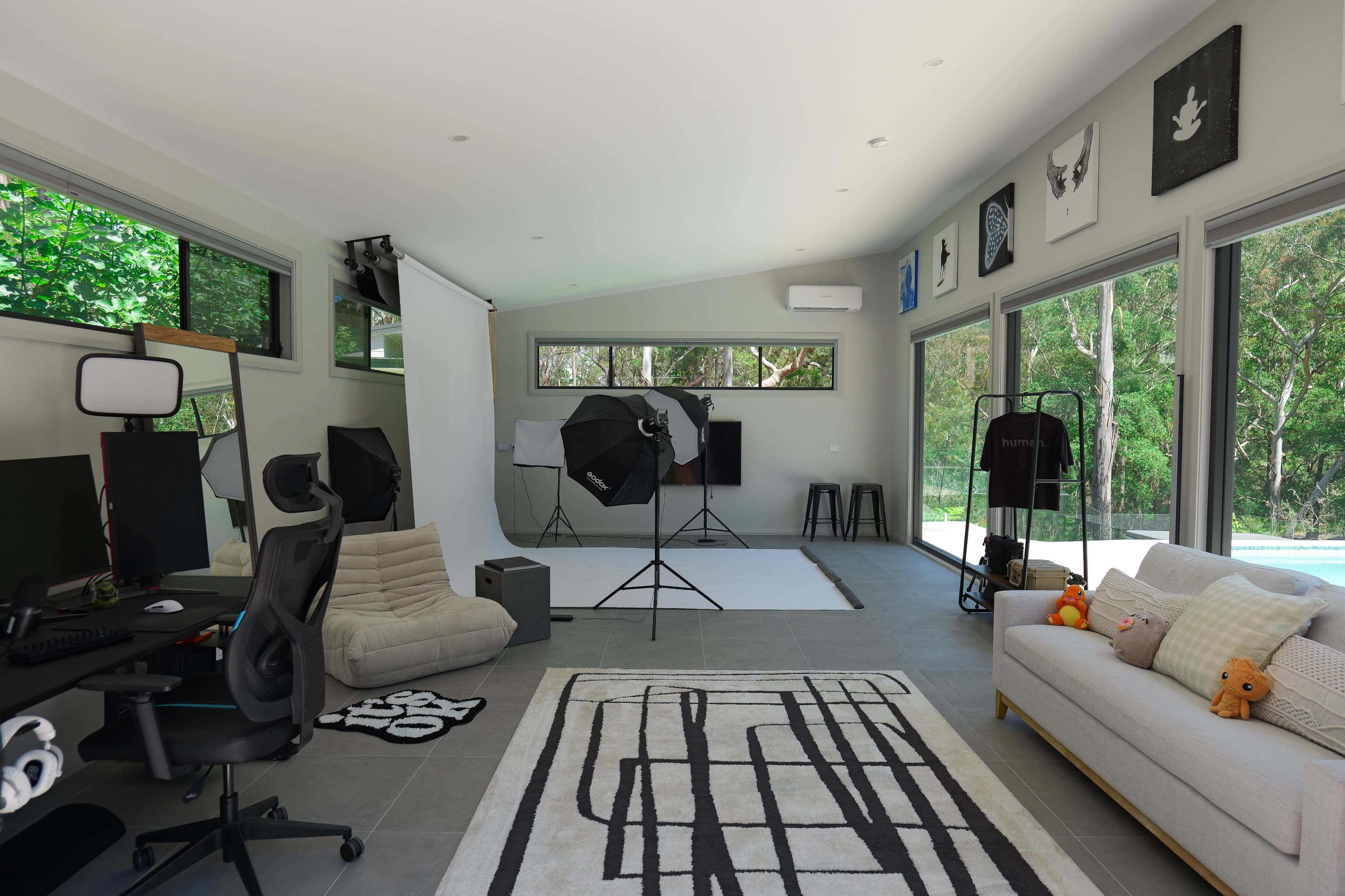 The image shows a modern studio space featuring a computer workstation, photography equipment, and large windows overlooking a green landscape.
