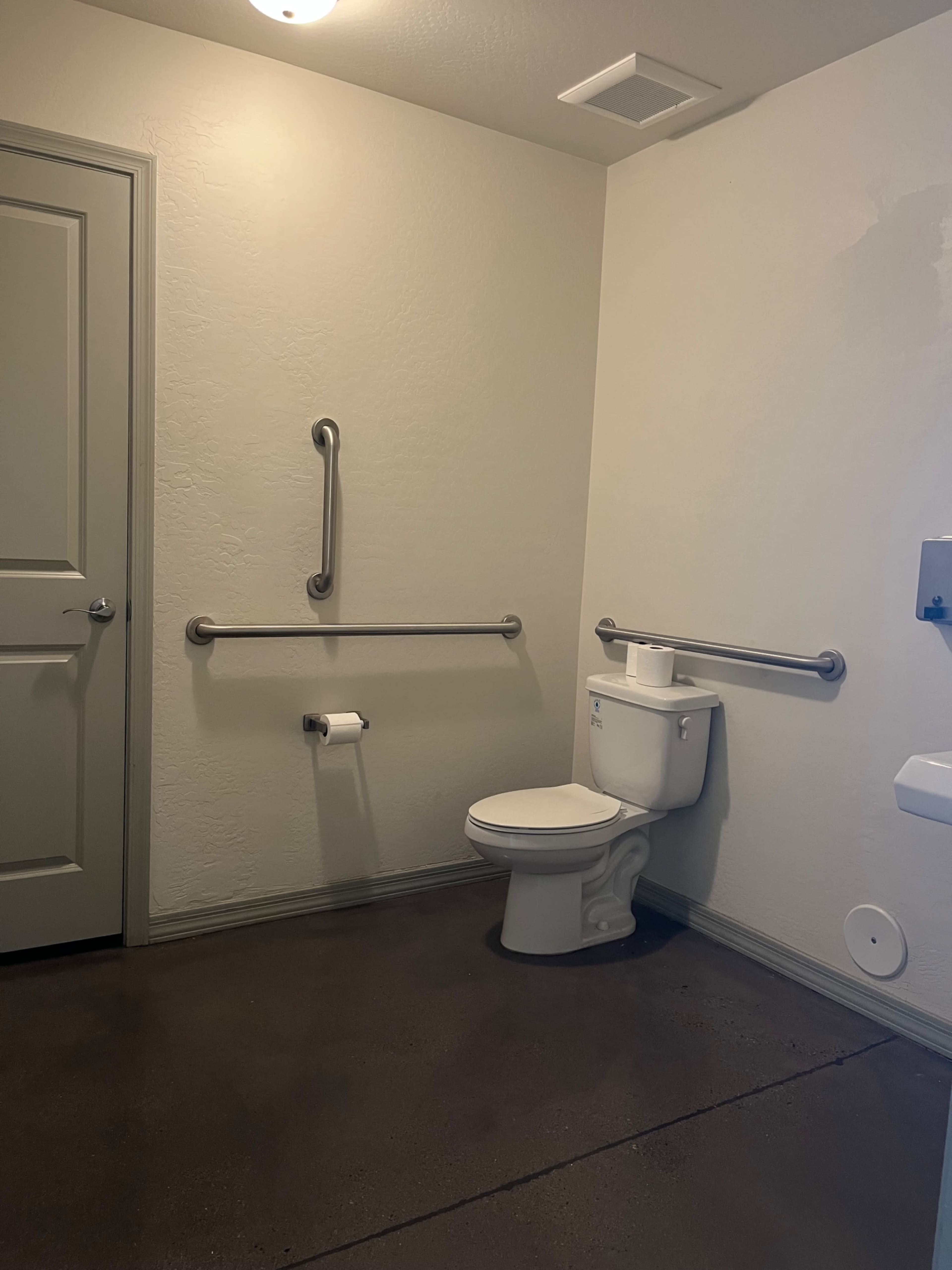 The image shows a restroom with a toilet, grab bars on the walls, and a door leading to another room.