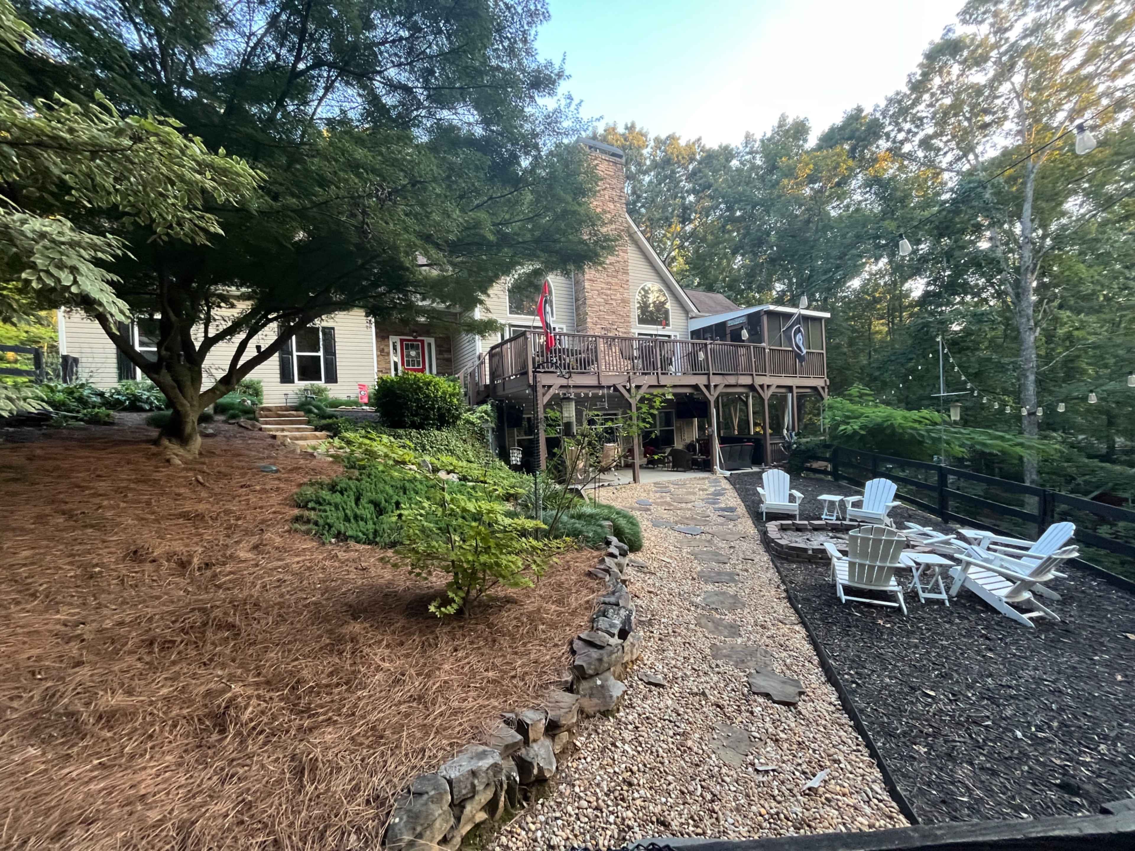 The image shows a house with a wooden deck, surrounded by landscaped greenery and a gravel pathway leading to a seating area with white adirondack chairs.