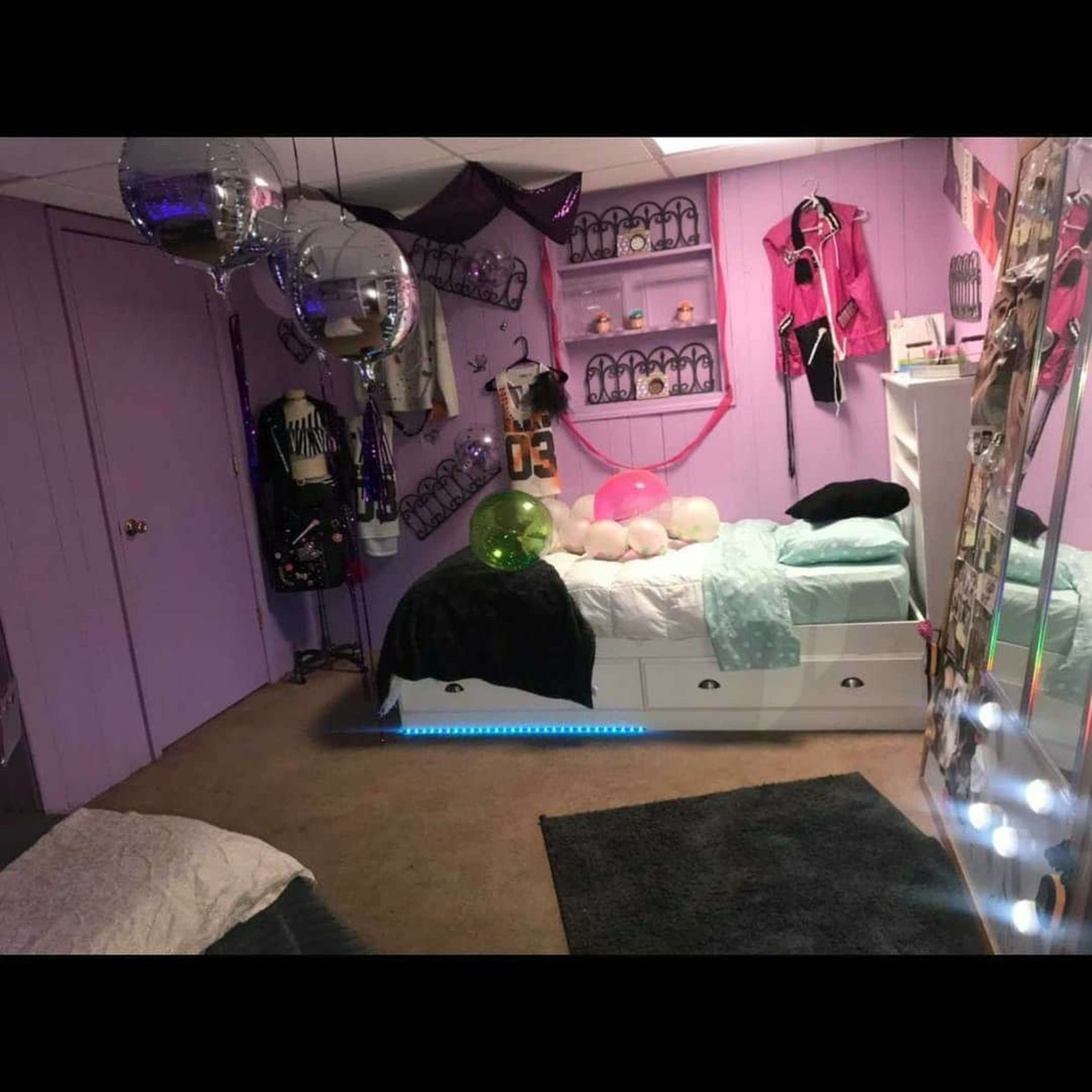 A purple-themed bedroom features a bed with colorful balloons, a mirror, and various decorations on the walls.