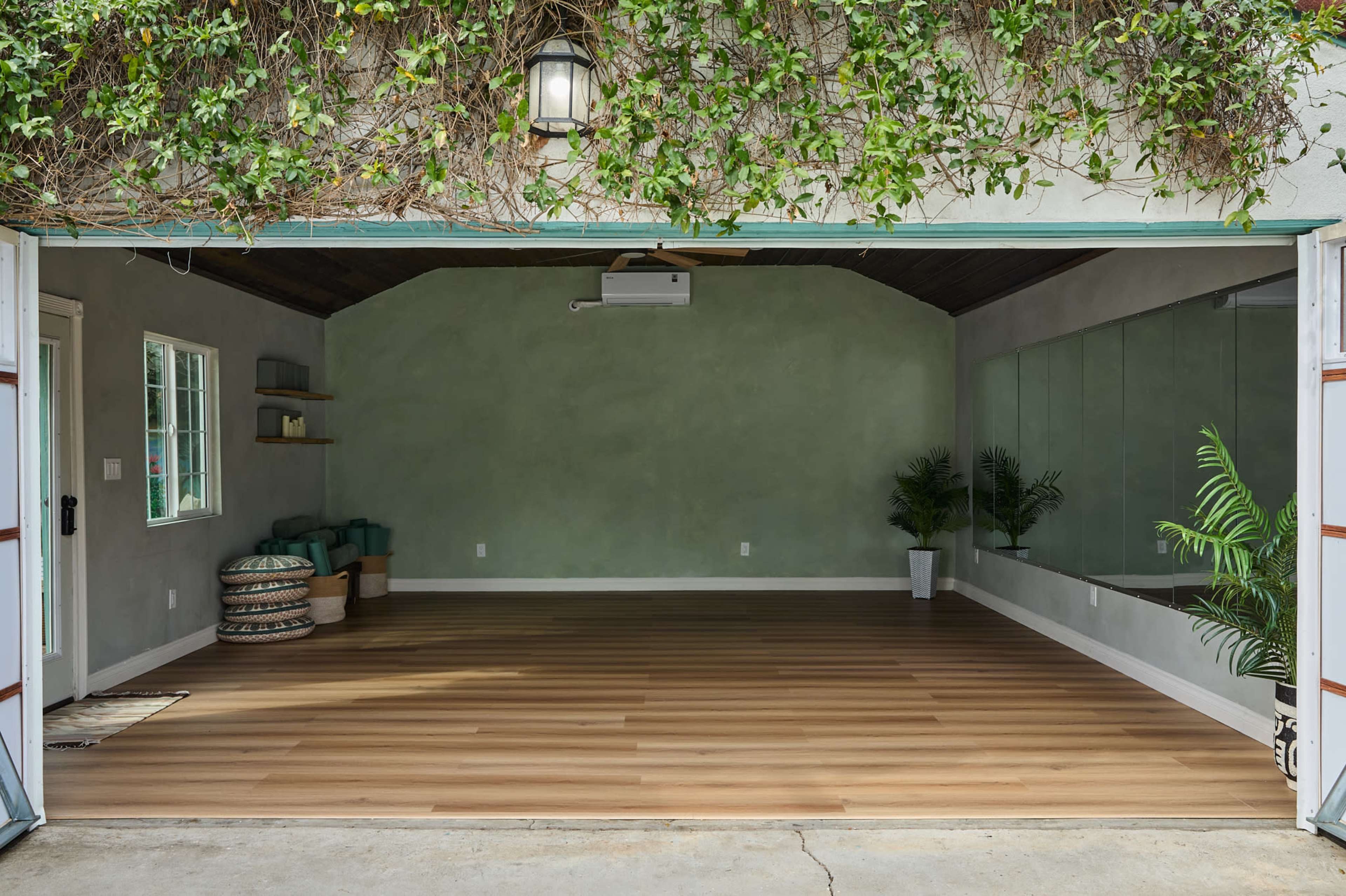 The image shows an empty, modern room with wooden flooring, a large mirror on one wall, and green plants in the corners, all under a ceiling with a light fixture and exposed beams.