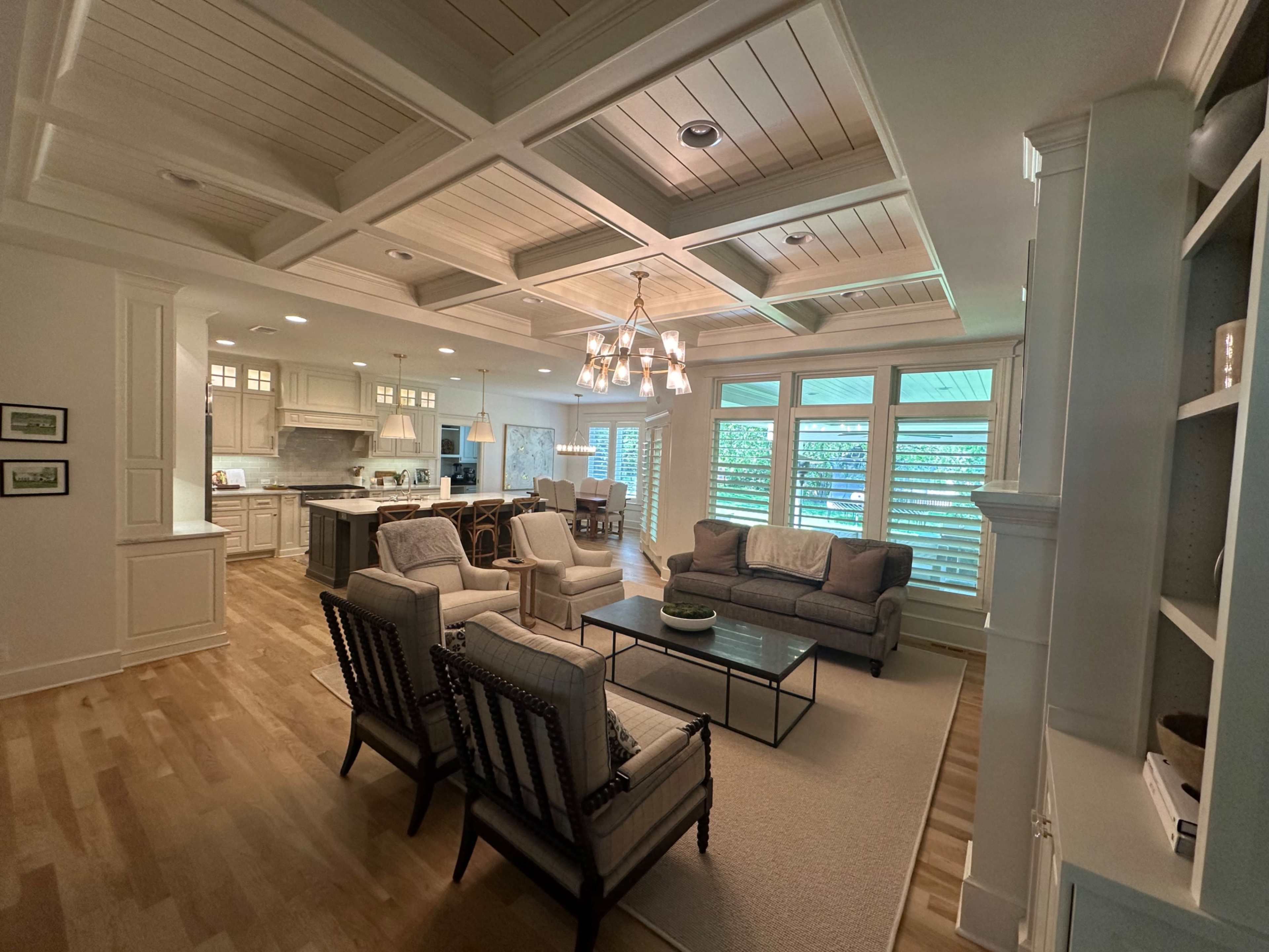 A spacious living room with a coffered ceiling, modern furniture, and large windows covered with plantation shutters.
