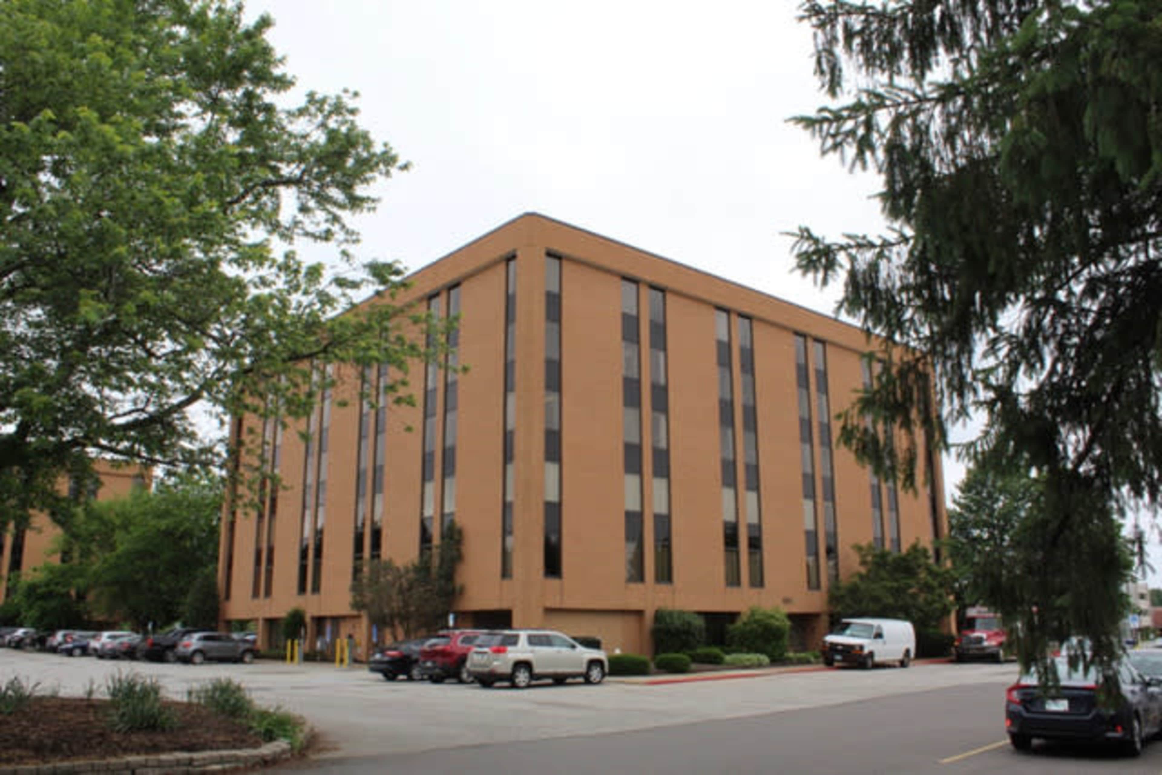 A four-story brick building with large windows is surrounded by trees and a parking lot filled with vehicles.