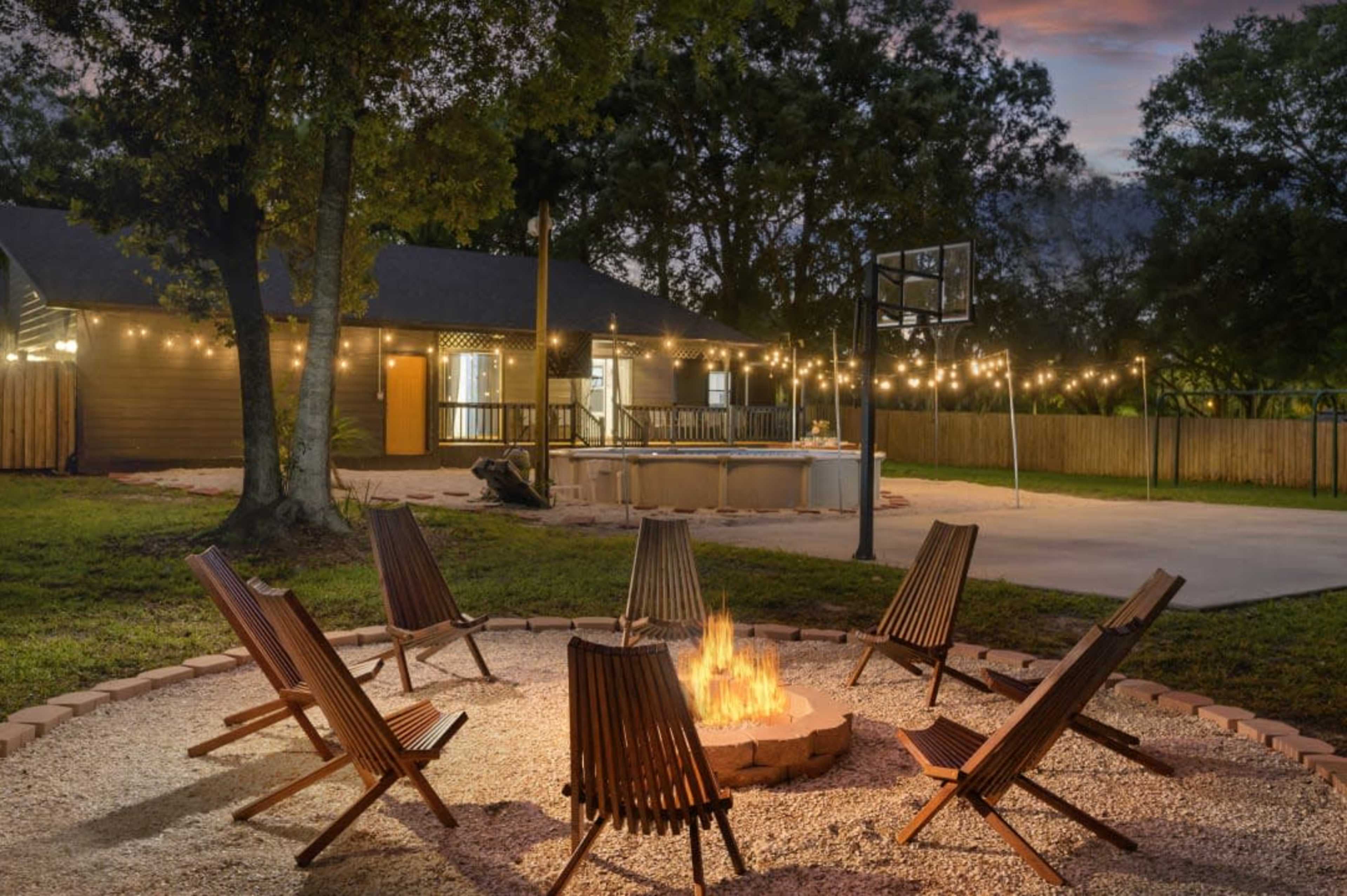 A circular fire pit surrounded by wooden chairs is situated in a backyard with string lights, a basketball hoop, and a house in the background.