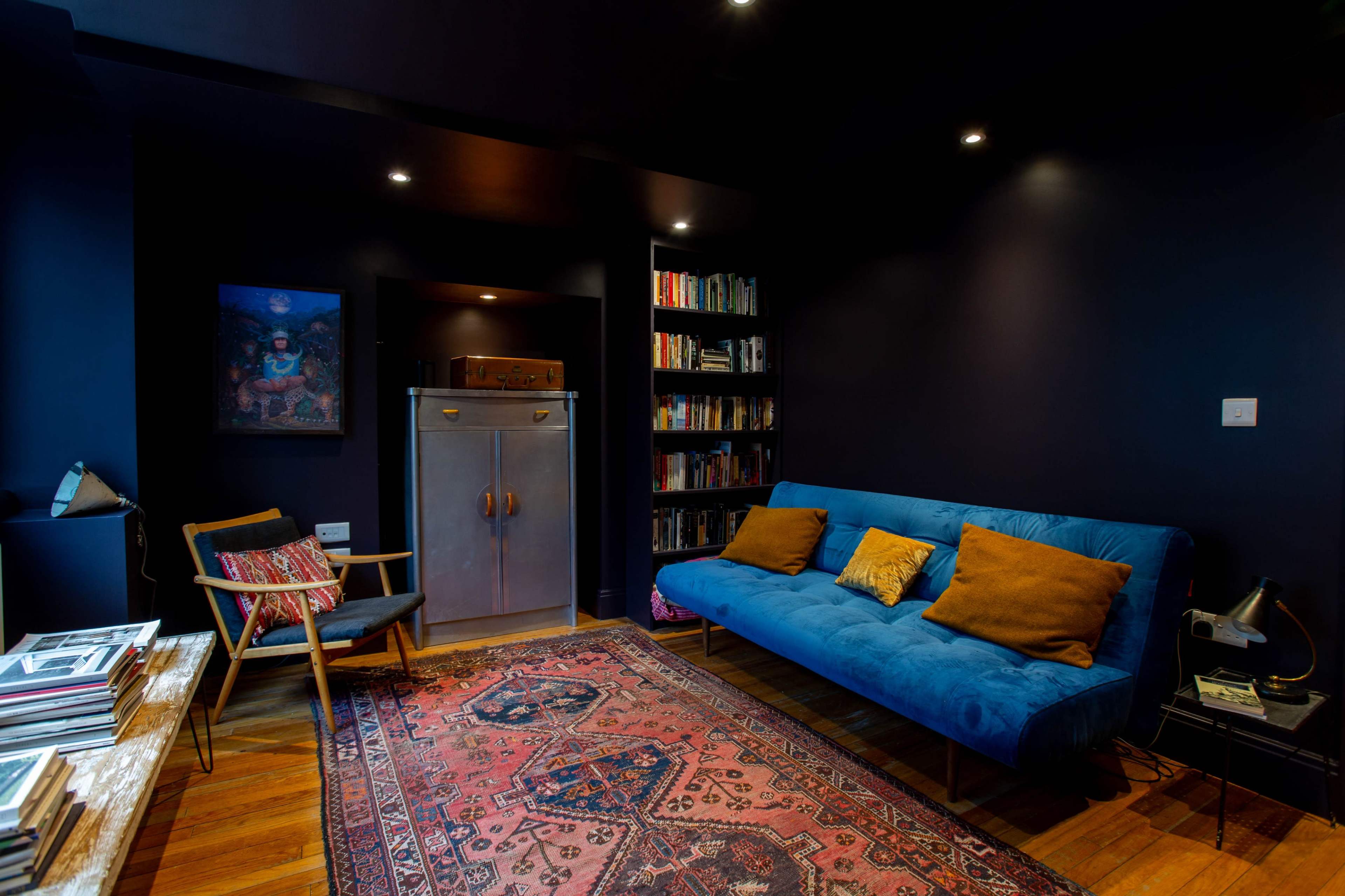 A dimly lit room features a blue sofa with decorative pillows, a wooden armchair, a bookshelf filled with books, and an antique cabinet against a dark wall.