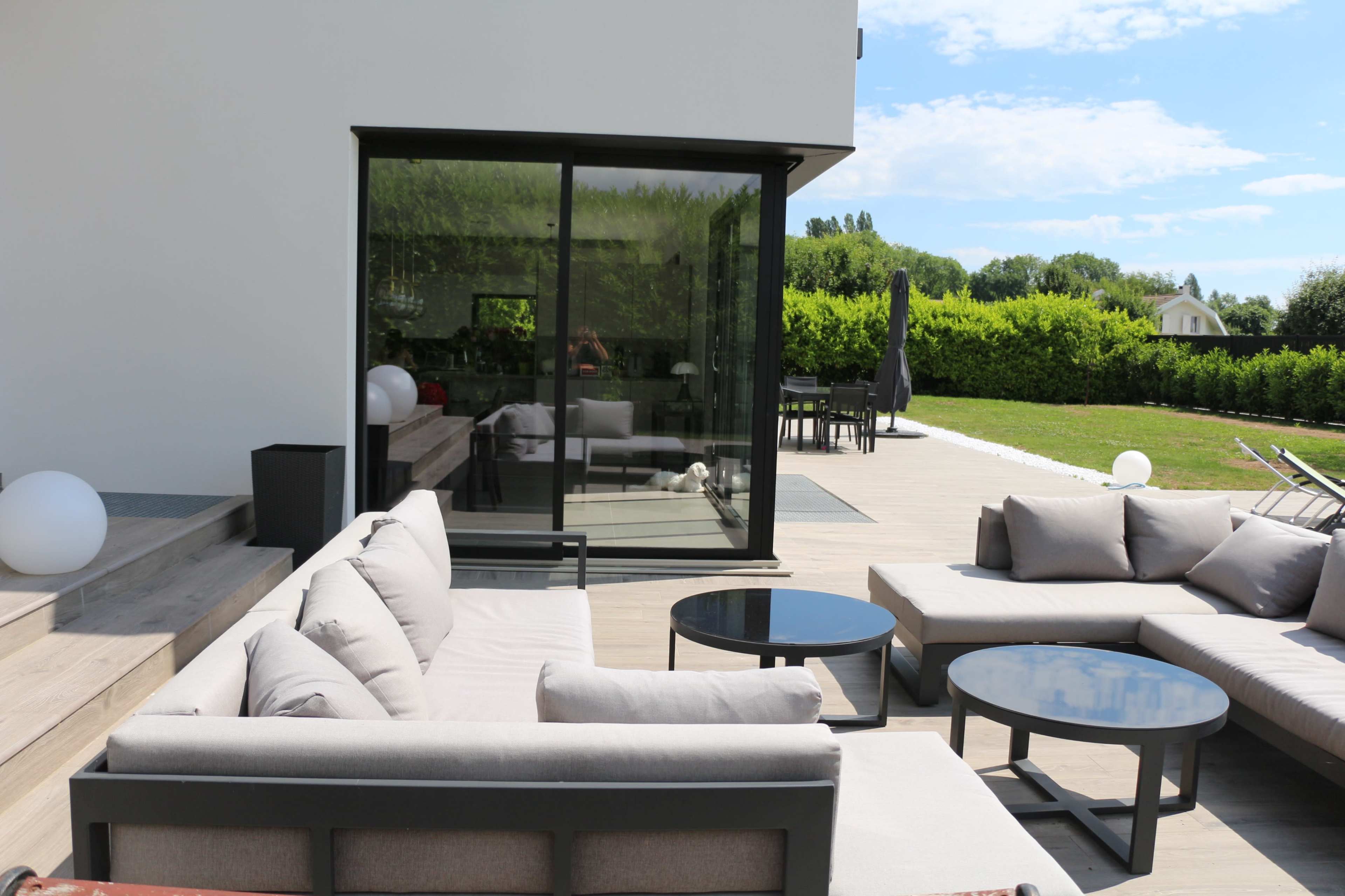 The image shows a modern outdoor patio area with gray seating, black tables, and a glass door leading to a green lawn.