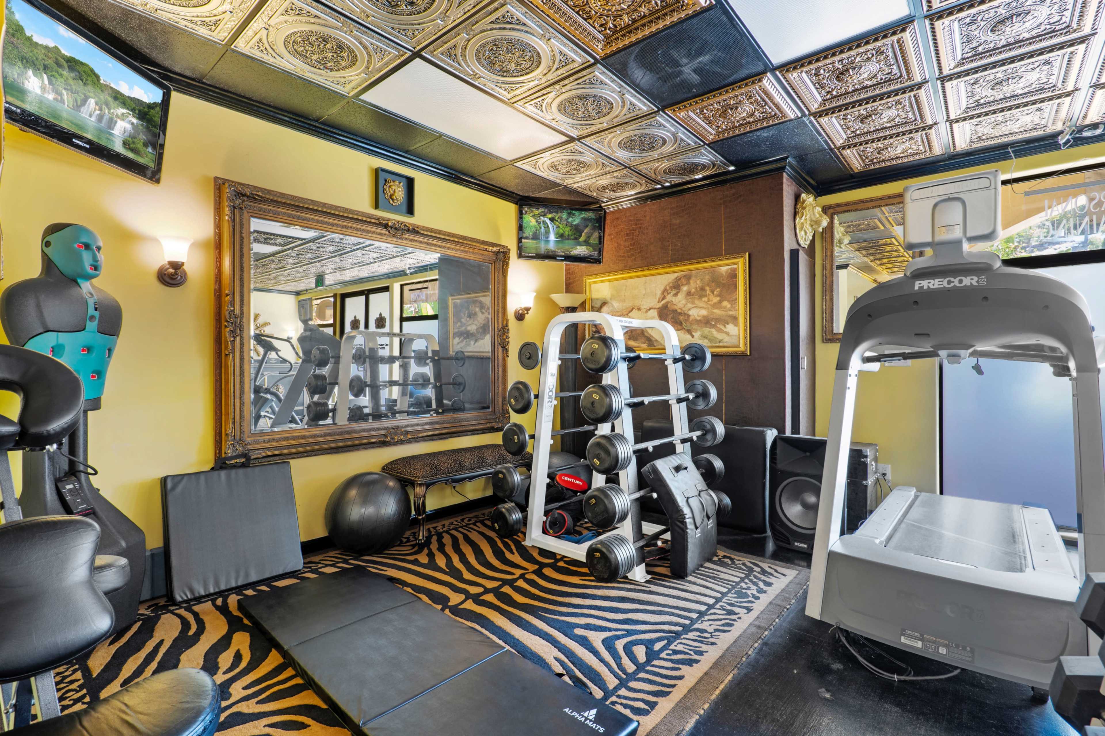 A home gym equipped with weightlifting equipment, a treadmill, and exercise mats, featuring a patterned rug and wall-mounted televisions.