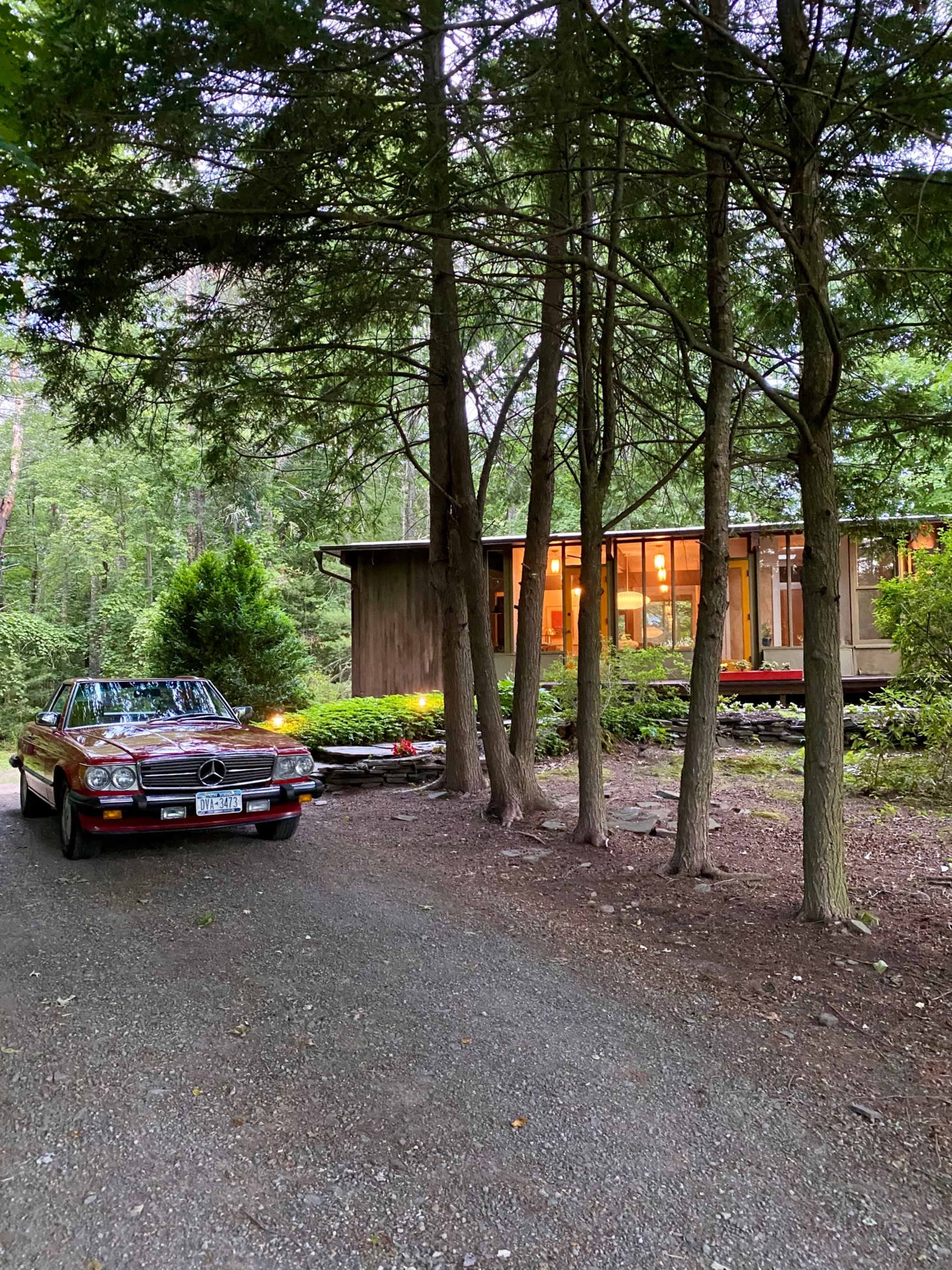 A red Mercedes car is parked on a gravel driveway beside a wooden house nestled among tall trees.