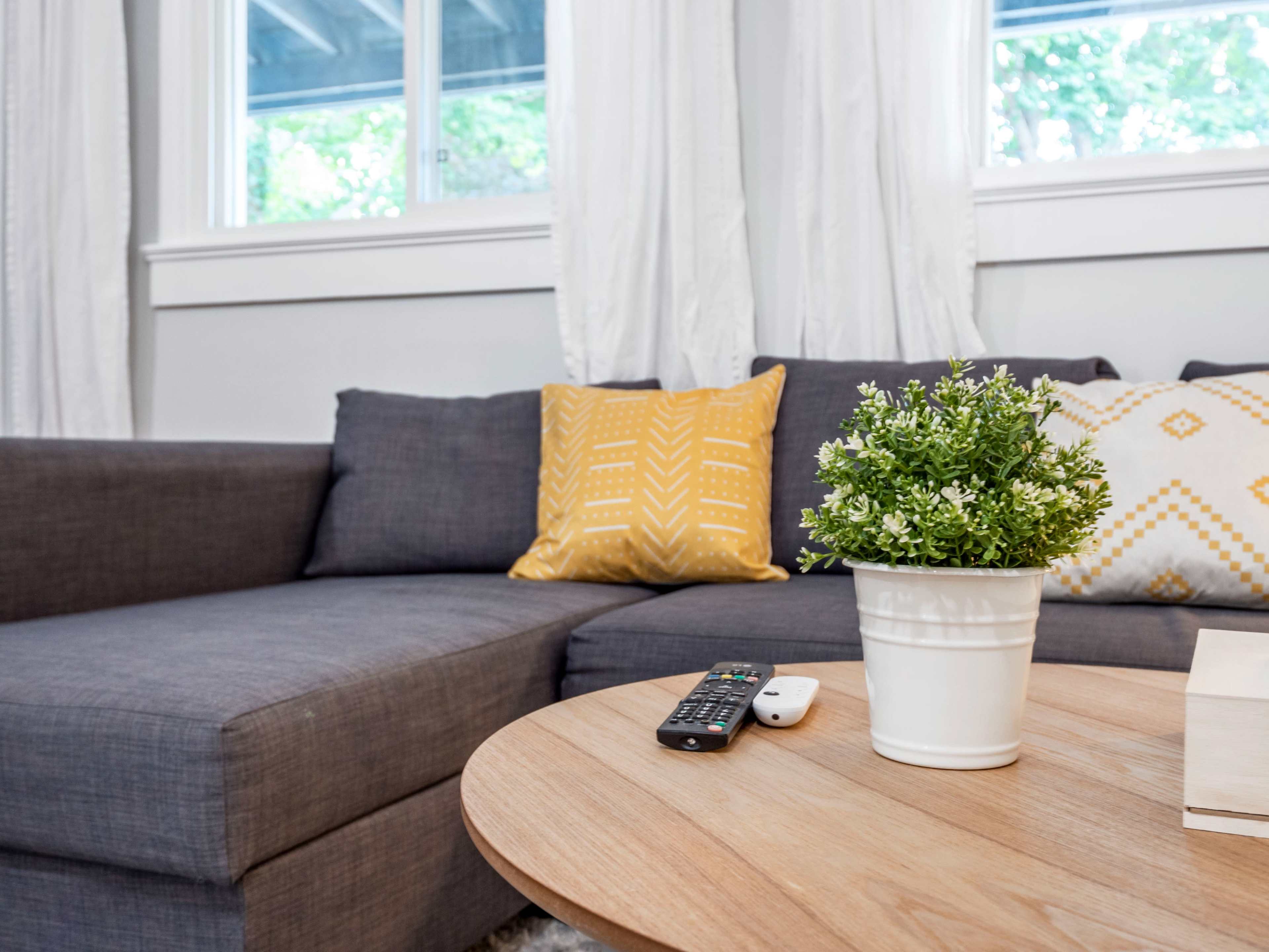 A gray sectional sofa with decorative yellow and white pillows sits beside a wooden coffee table featuring a small potted plant and a remote control.