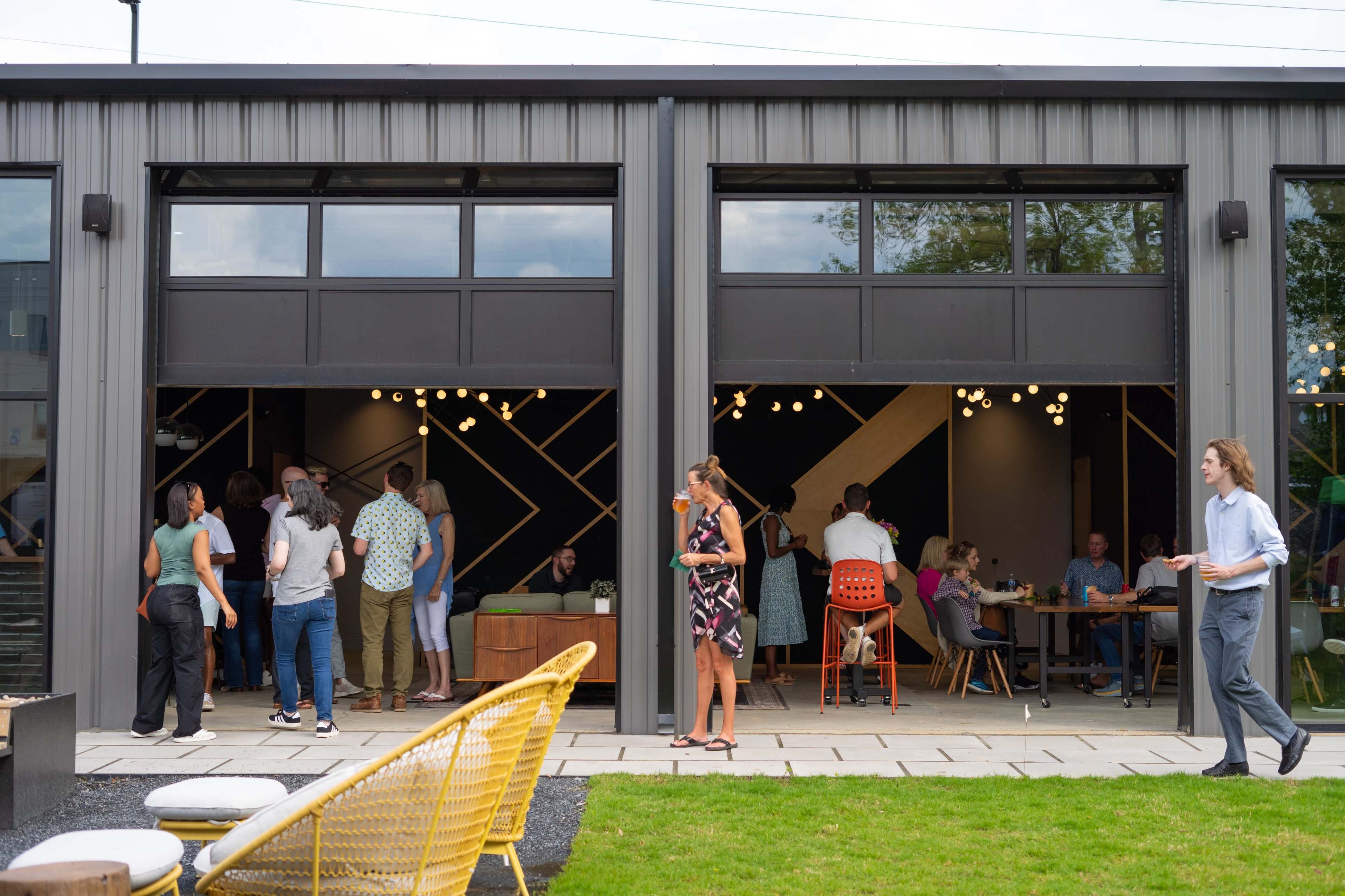 A group of people socializes and works inside and outside a modern building with large open doors, showcasing a lively atmosphere.