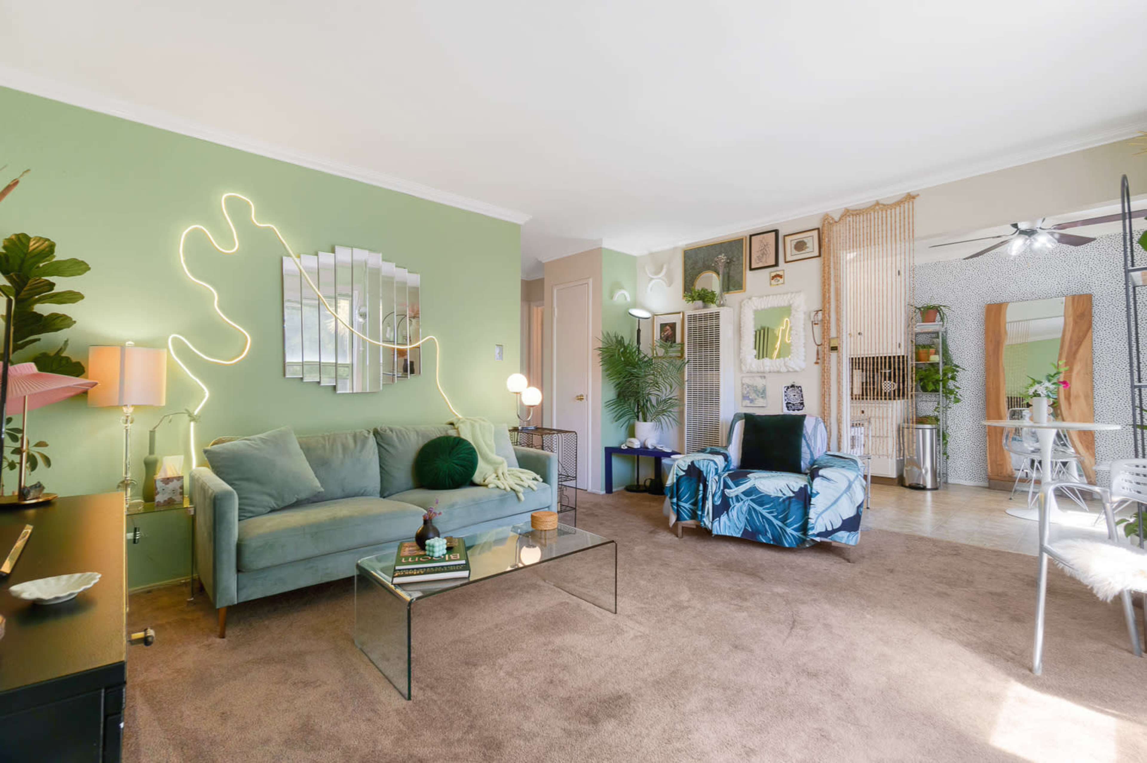 The living room features a green wall, a velvet sofa, a glass coffee table, and a decorated space with plants and framed art.