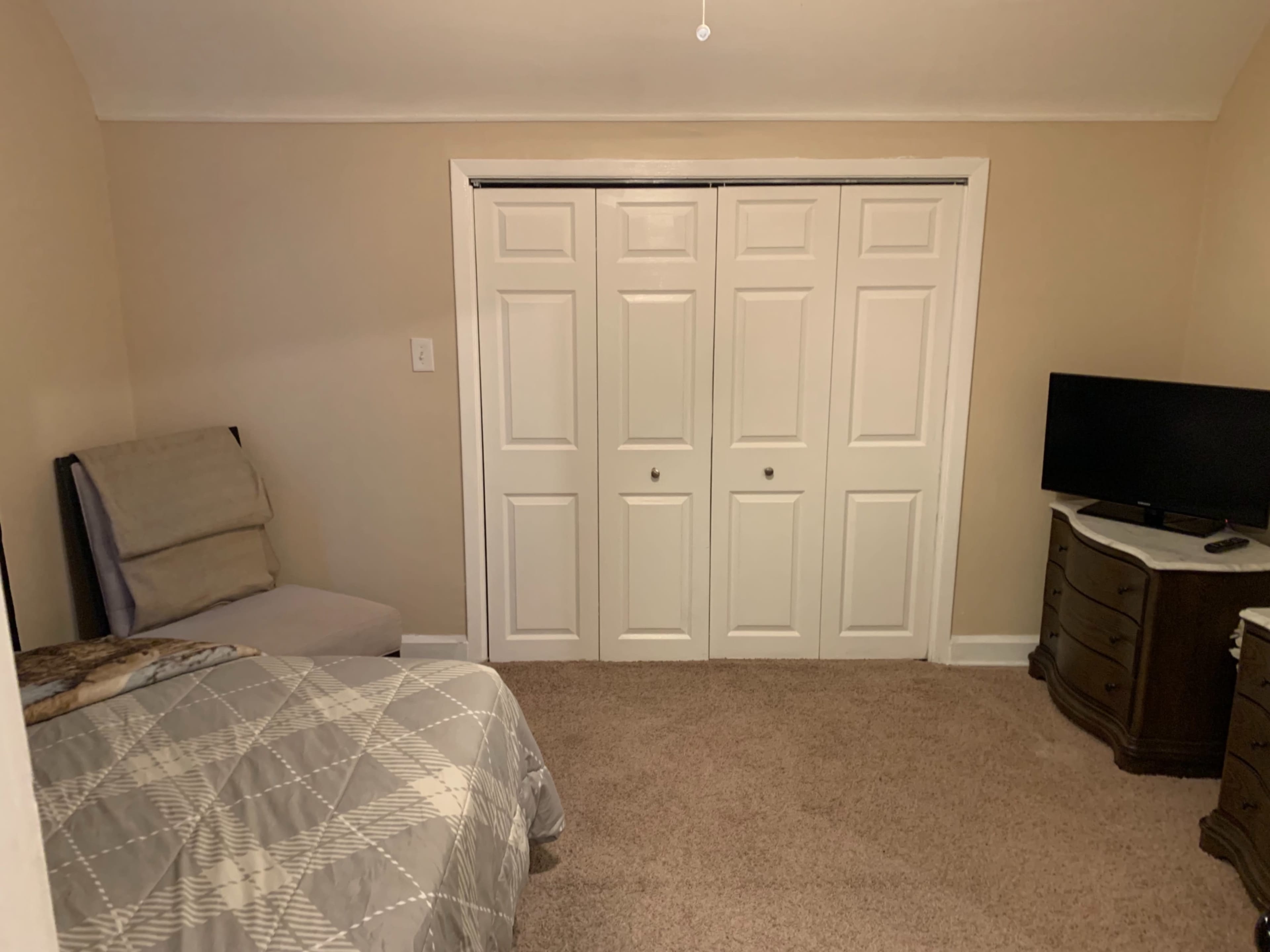 The image shows a small bedroom with a bed covered in a patterned blanket, a chair, a TV on a dresser, and a closet with sliding doors.