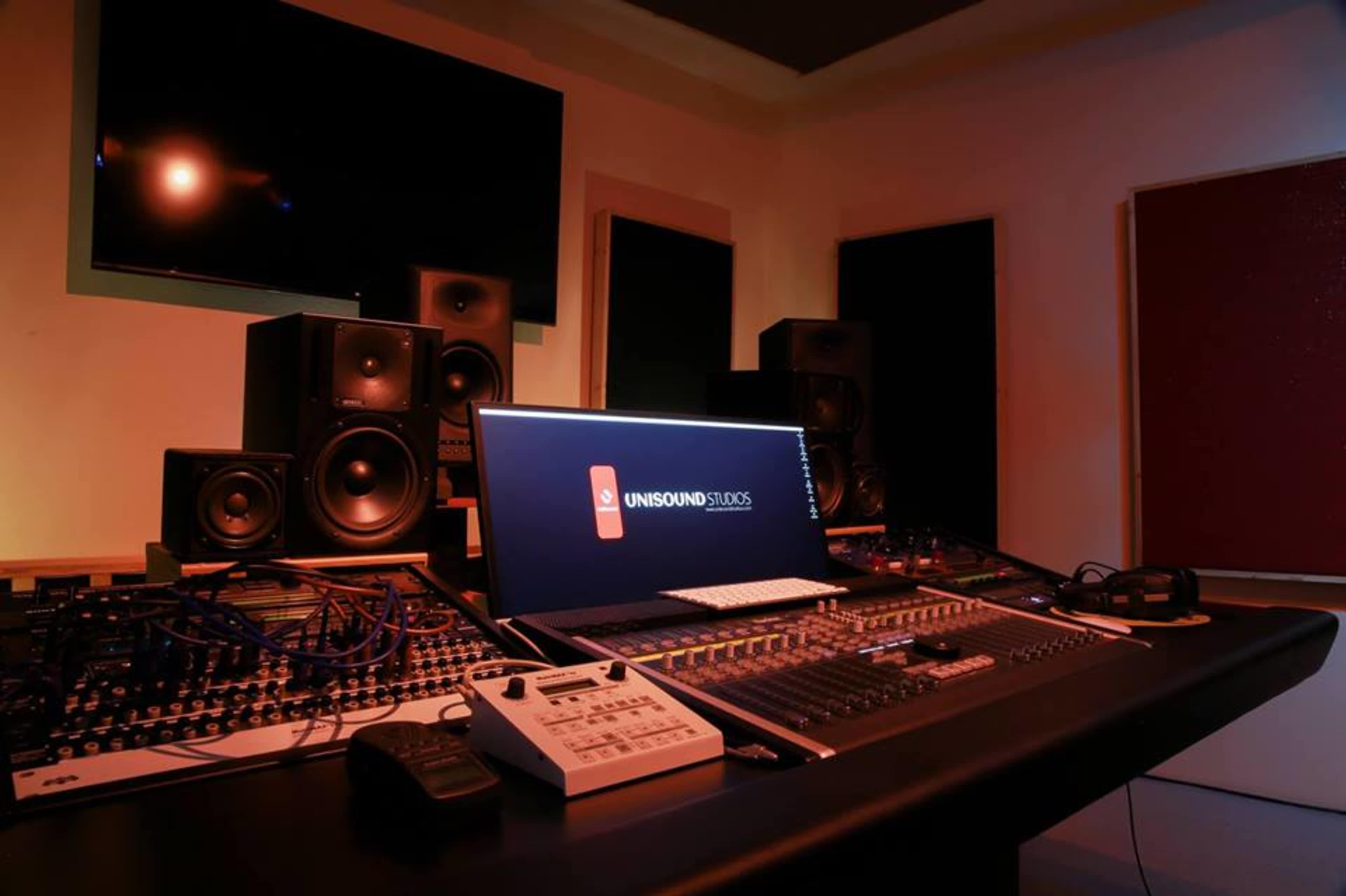 A music production studio with a mixing console, computer display, and several audio speakers arranged around a dark table.