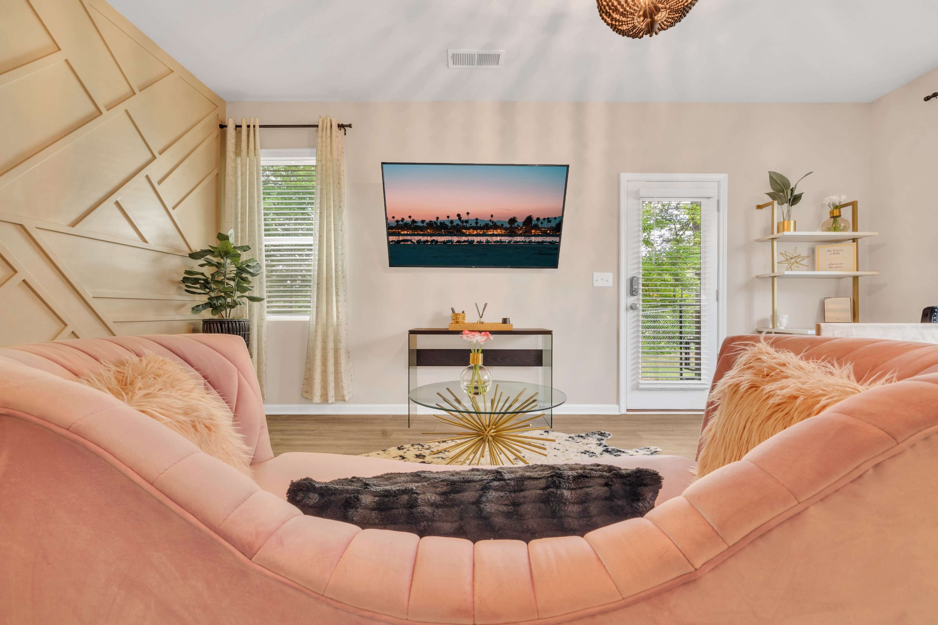 A modern living room features a pink velvet sofa, a glass coffee table with a gold base, and a wall-mounted television displaying a sunset scene.