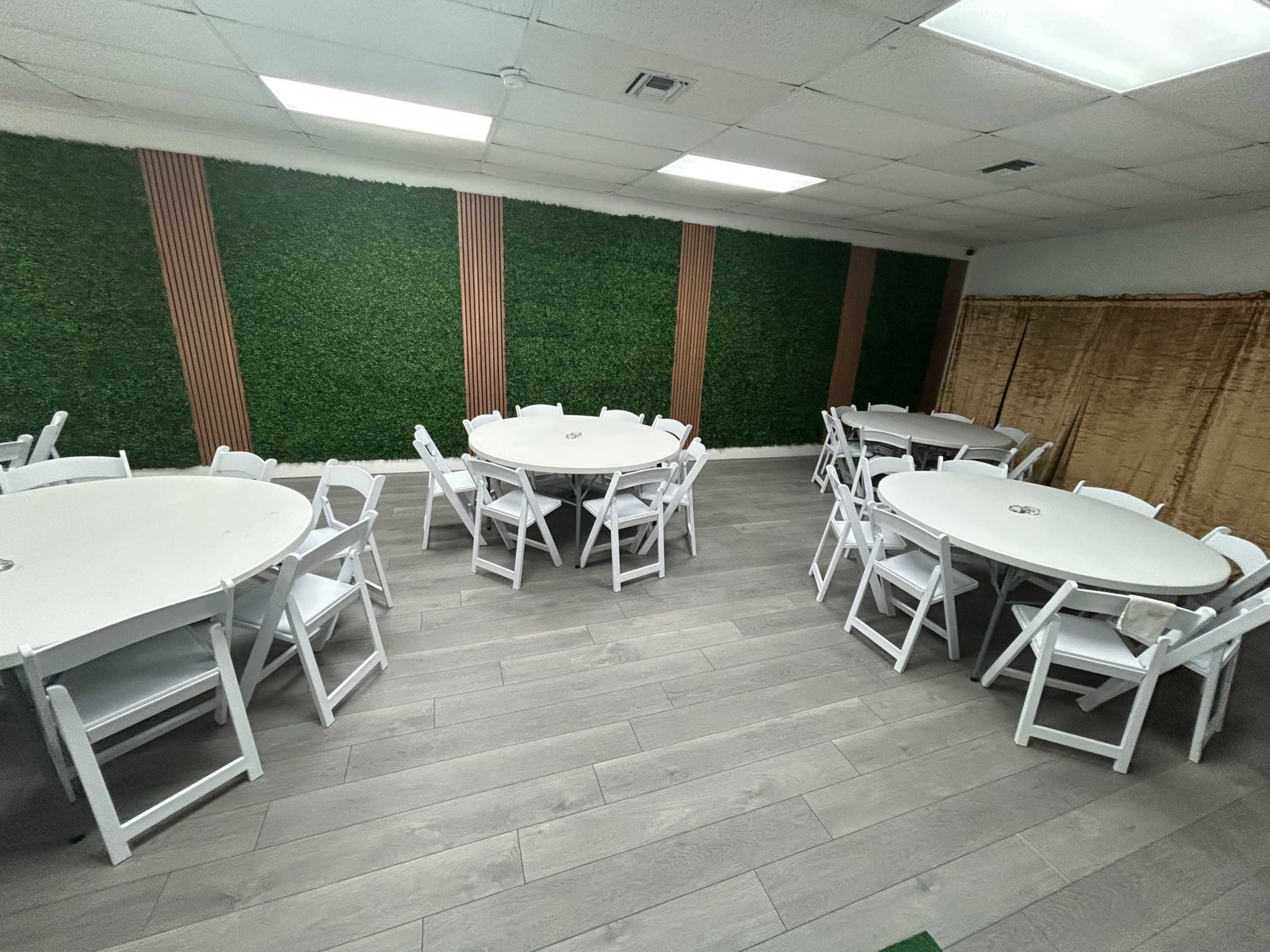 The image shows a room with several round tables surrounded by white chairs, set against a backdrop of green wall foliage.