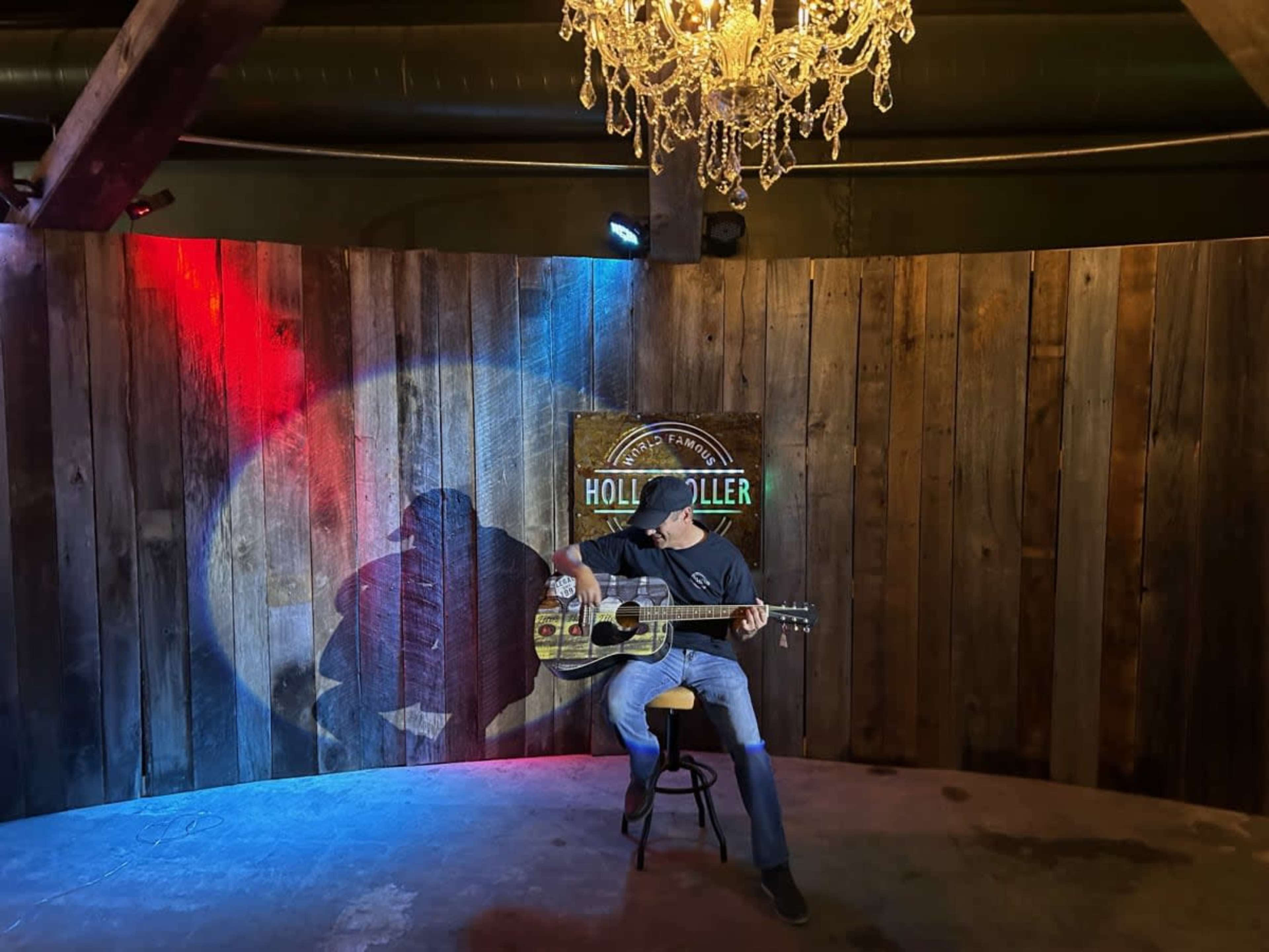 A musician sits on a stool playing an acoustic guitar in front of a wooden backdrop with a chandelier overhead and colored lights casting shadows.
