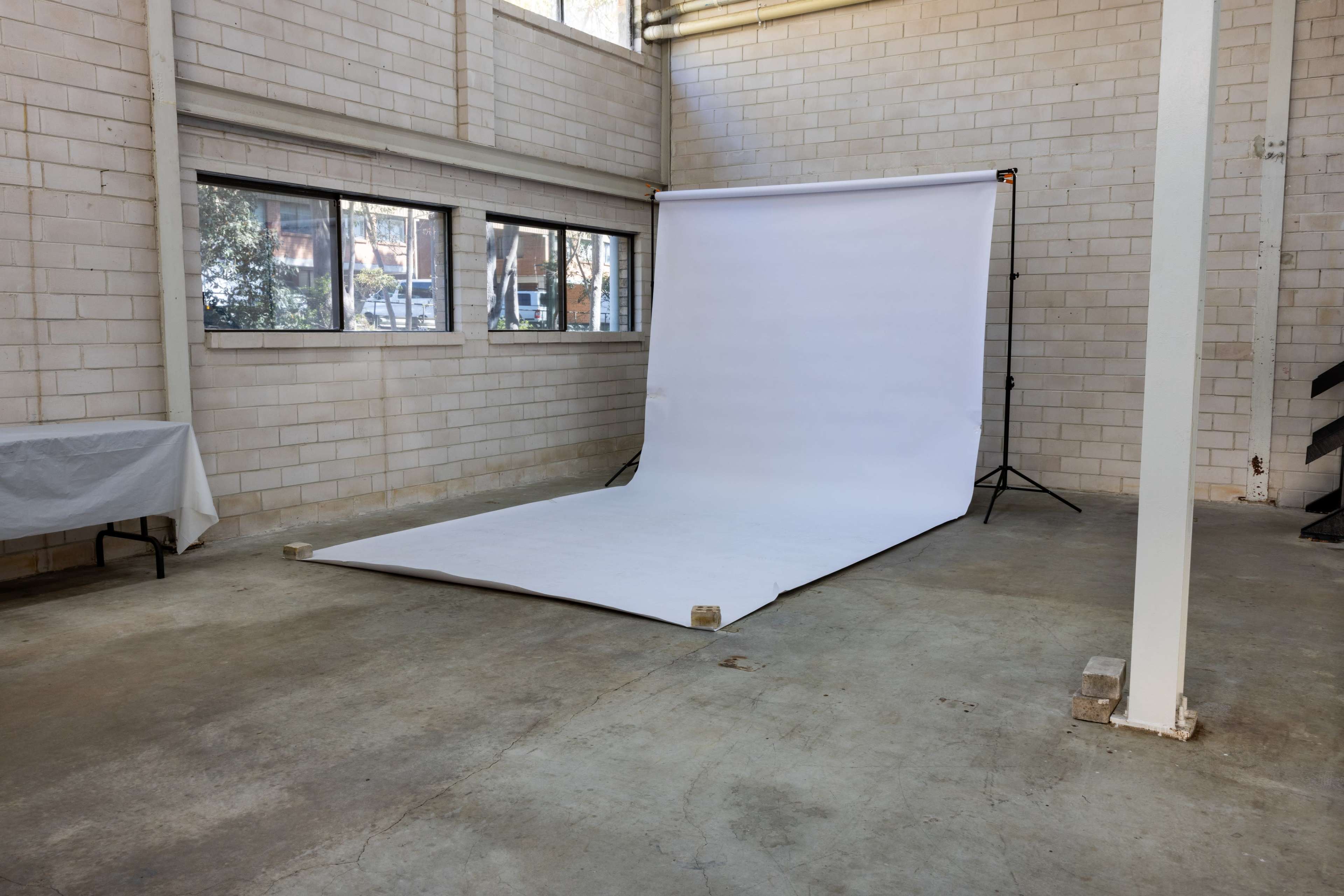 A photography studio features a white backdrop set up on the floor with large windows and bare concrete walls.