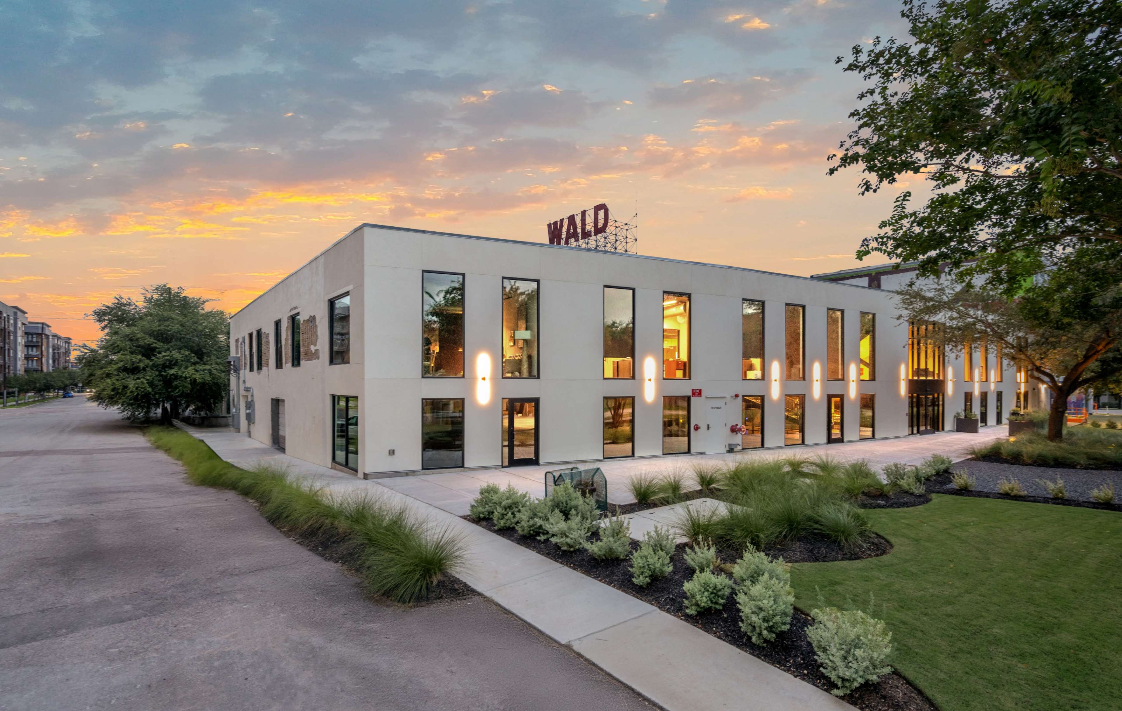 The image shows a modern building with large windows and a "WALD" sign, surrounded by landscaped grass and trees under a colorful sunset sky.