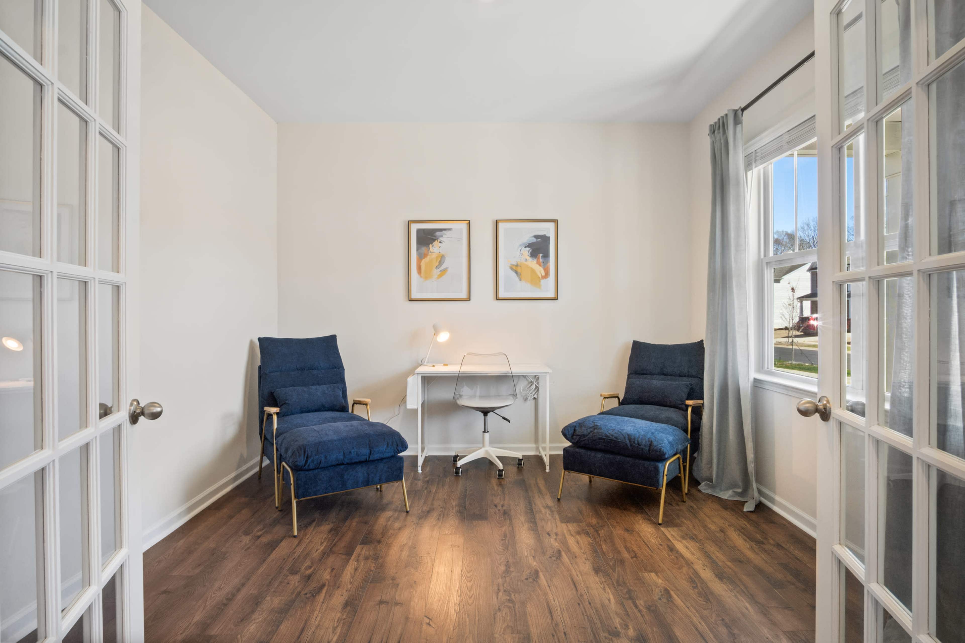 A bright room featuring two blue chairs, a small white desk with a lamp, and framed artwork on the walls.