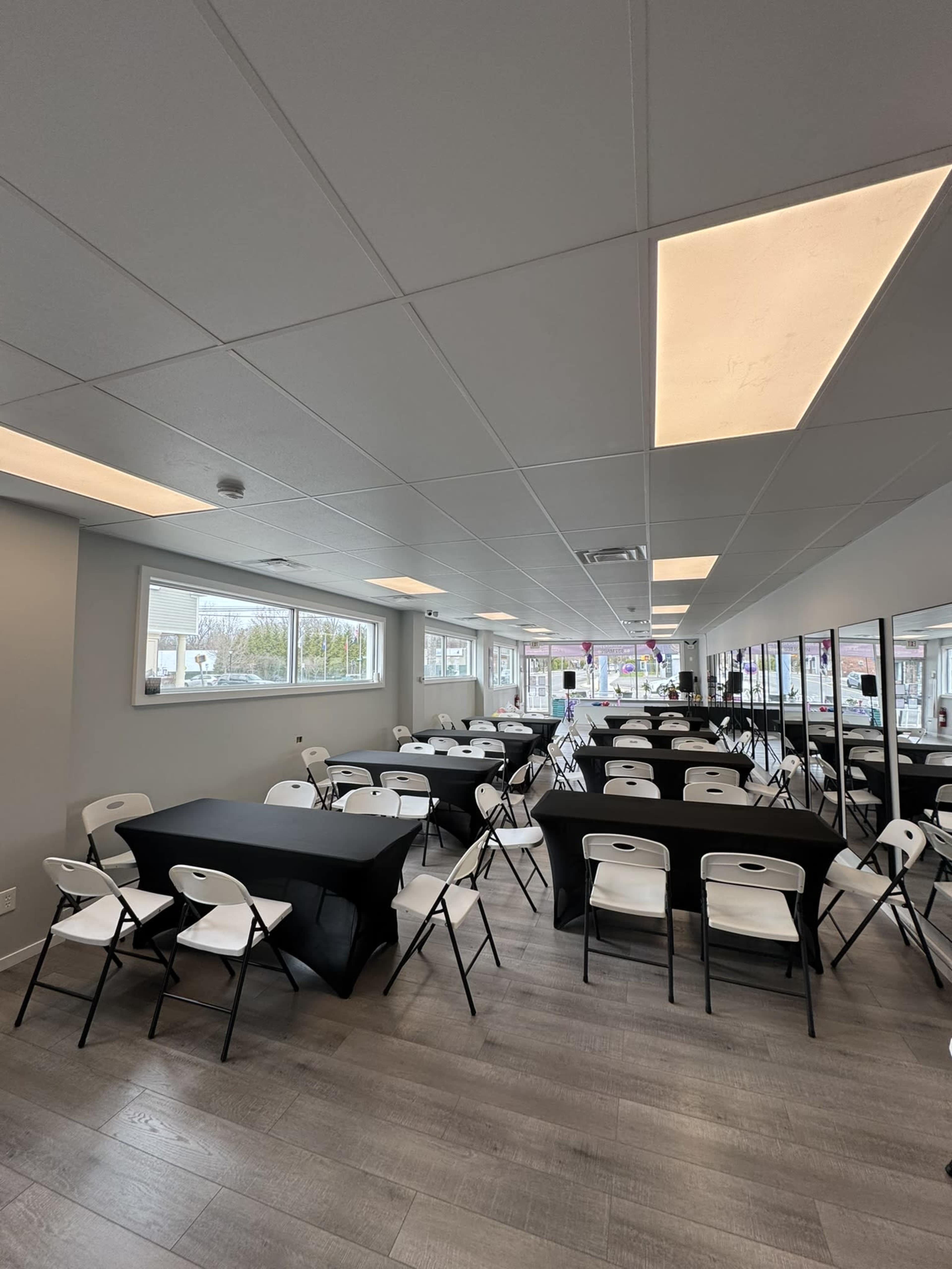 The image shows a spacious room set up for an event, featuring several black tables covered with tablecloths and surrounded by white folding chairs.