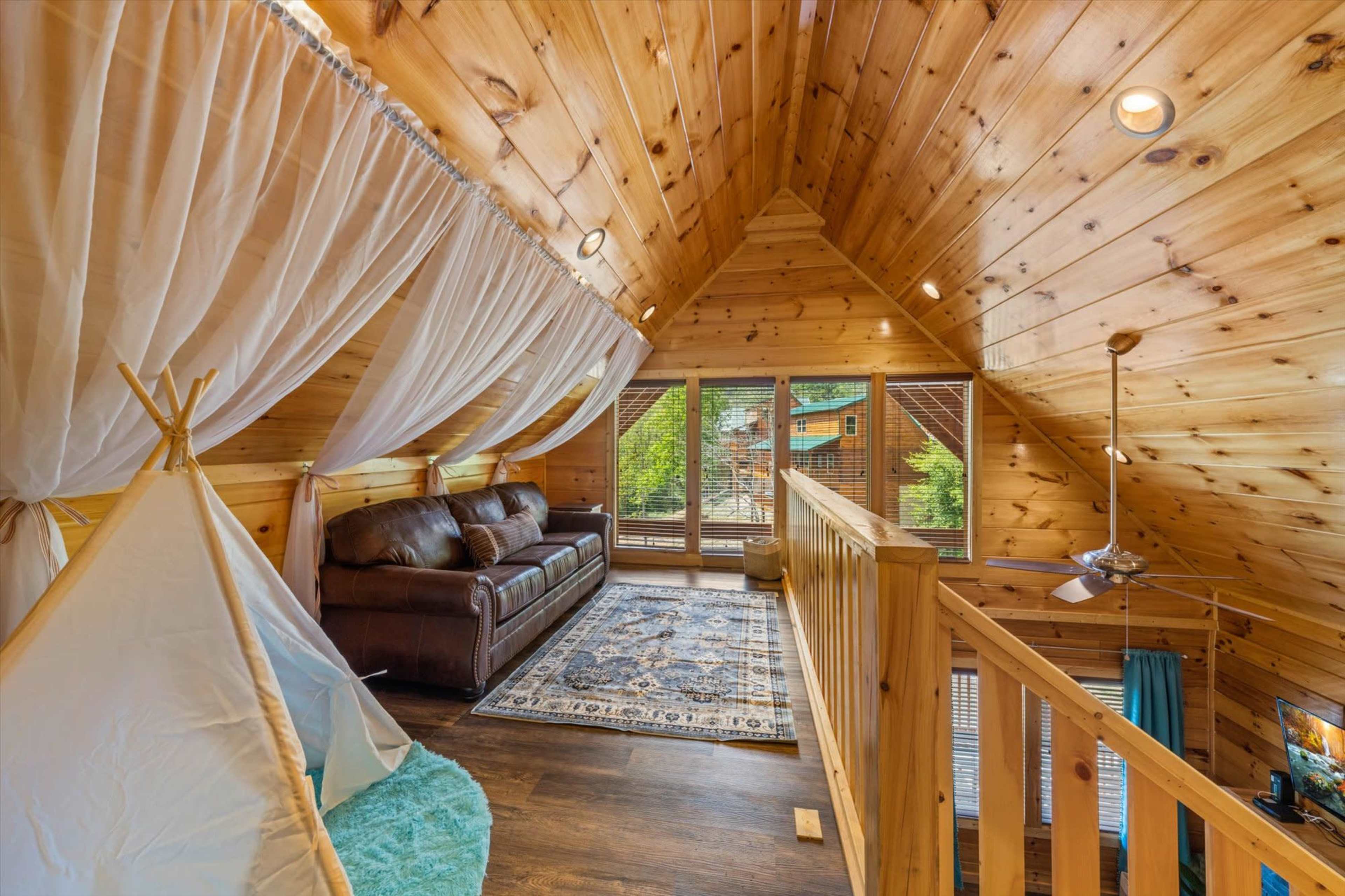 A cozy loft area with a wooden ceiling, a brown leather couch, a patterned rug, and a children's play tent.