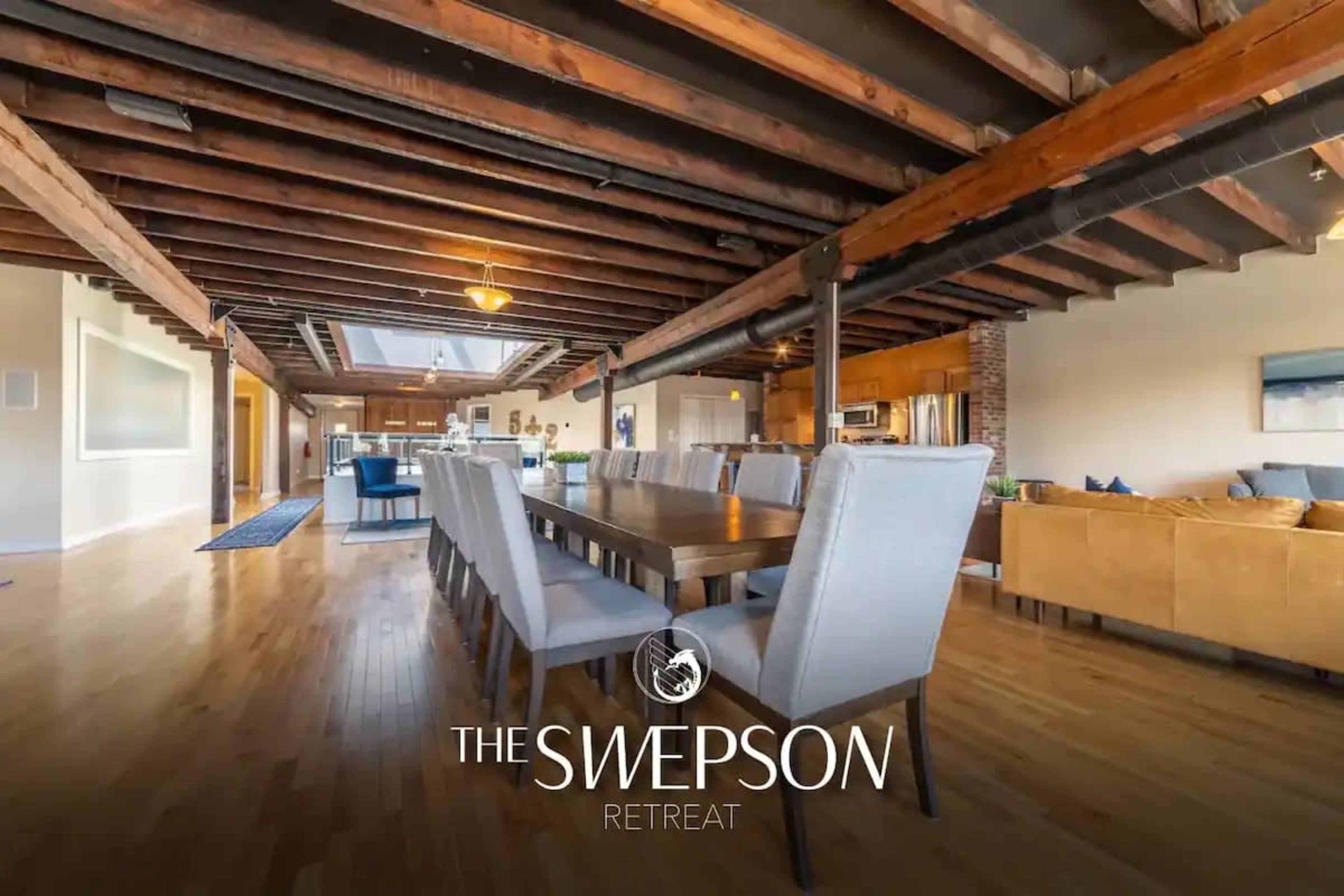 A spacious open-concept living area featuring a long dining table, wooden beams, and large windows that fill the room with natural light.