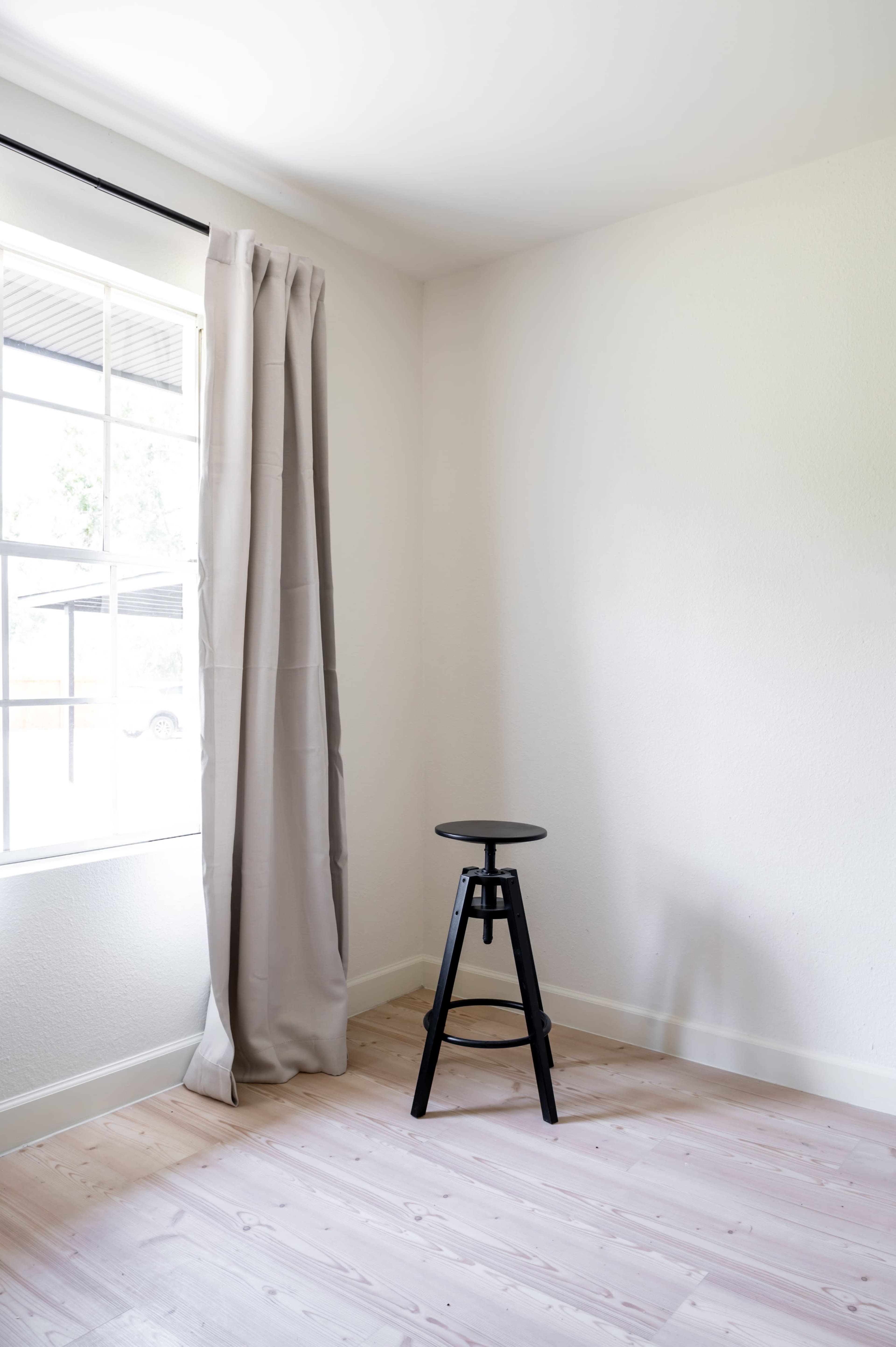 A black stool is positioned in a bright corner of a room with light-colored wooden flooring and a window covered by a beige curtain.
