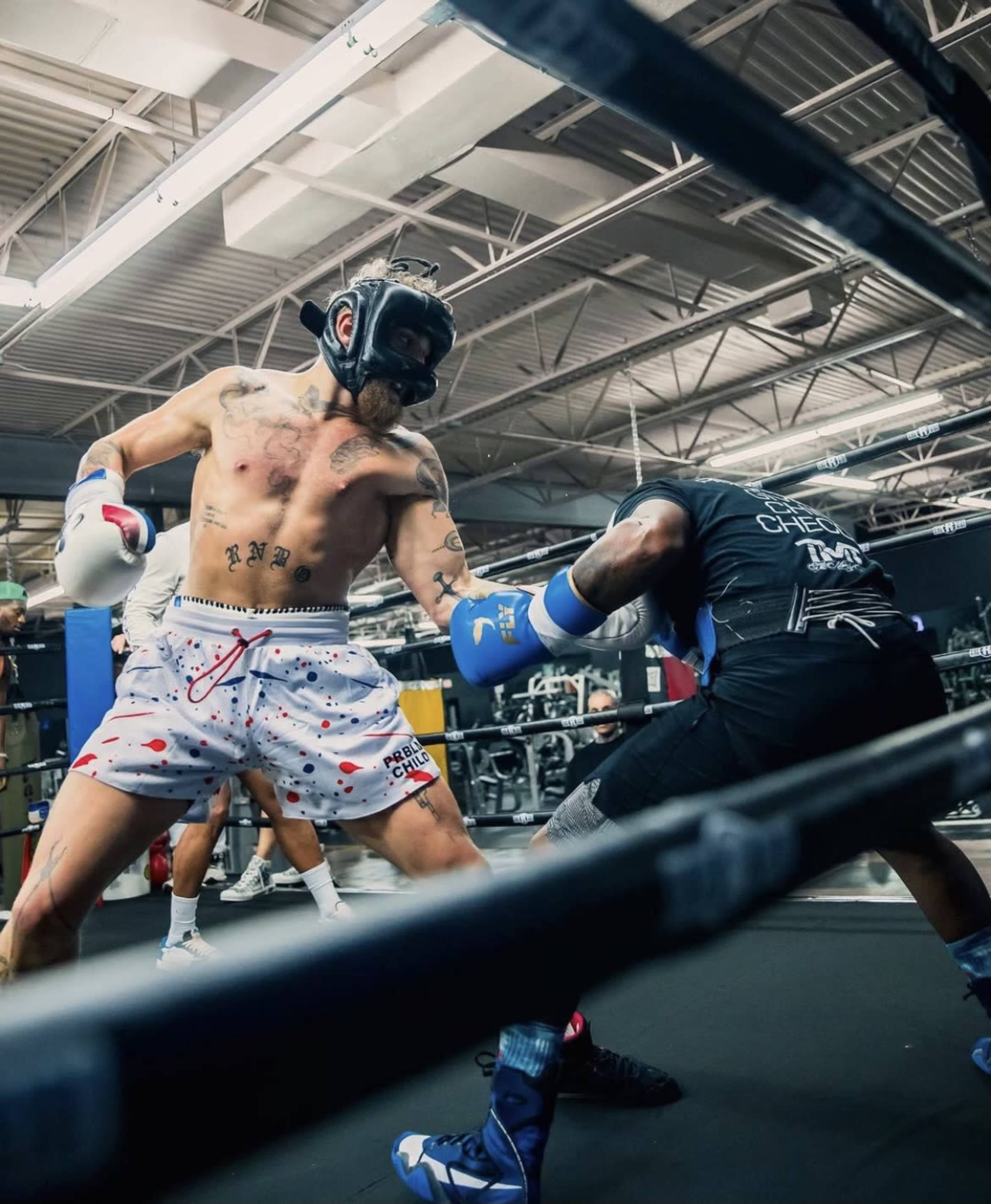 A boxer wearing blue gloves and a protective helmet throws a punch while sparring with another boxer in a gym setting.