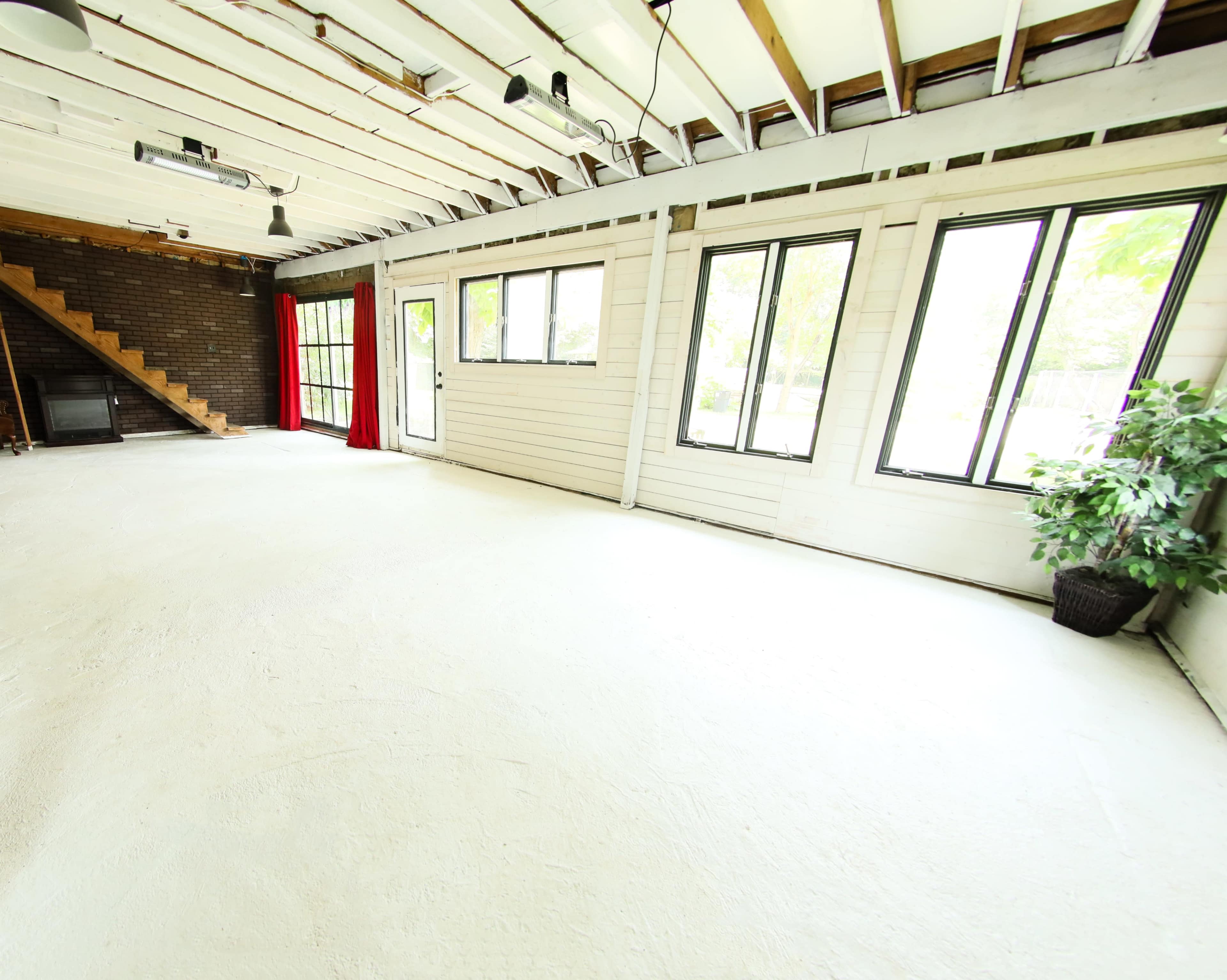 The image shows a spacious room with high ceilings, large windows, and a wooden staircase in the background.