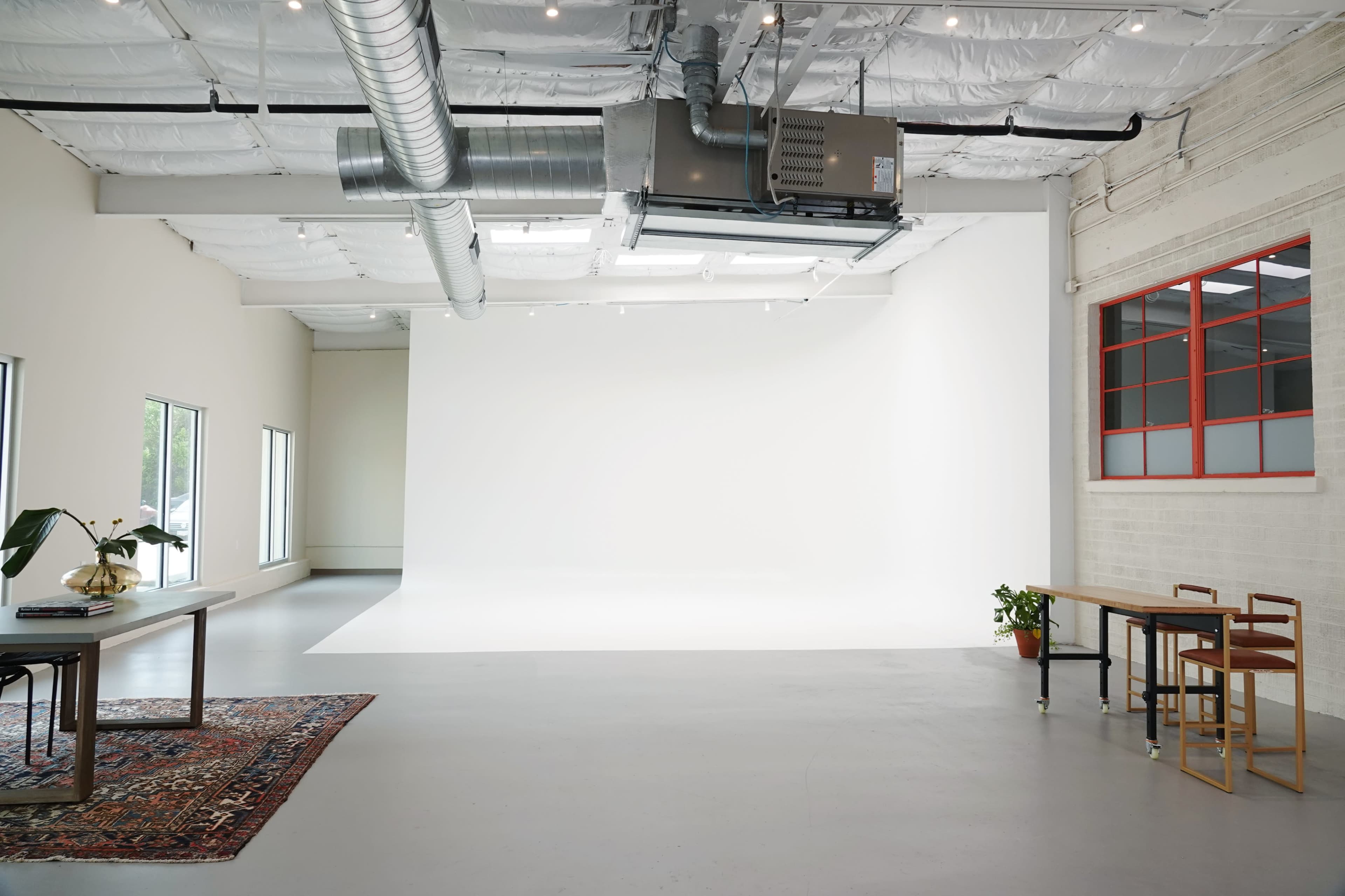 A spacious studio features a white backdrop, large windows, and minimalist furniture, including a table and a rug.