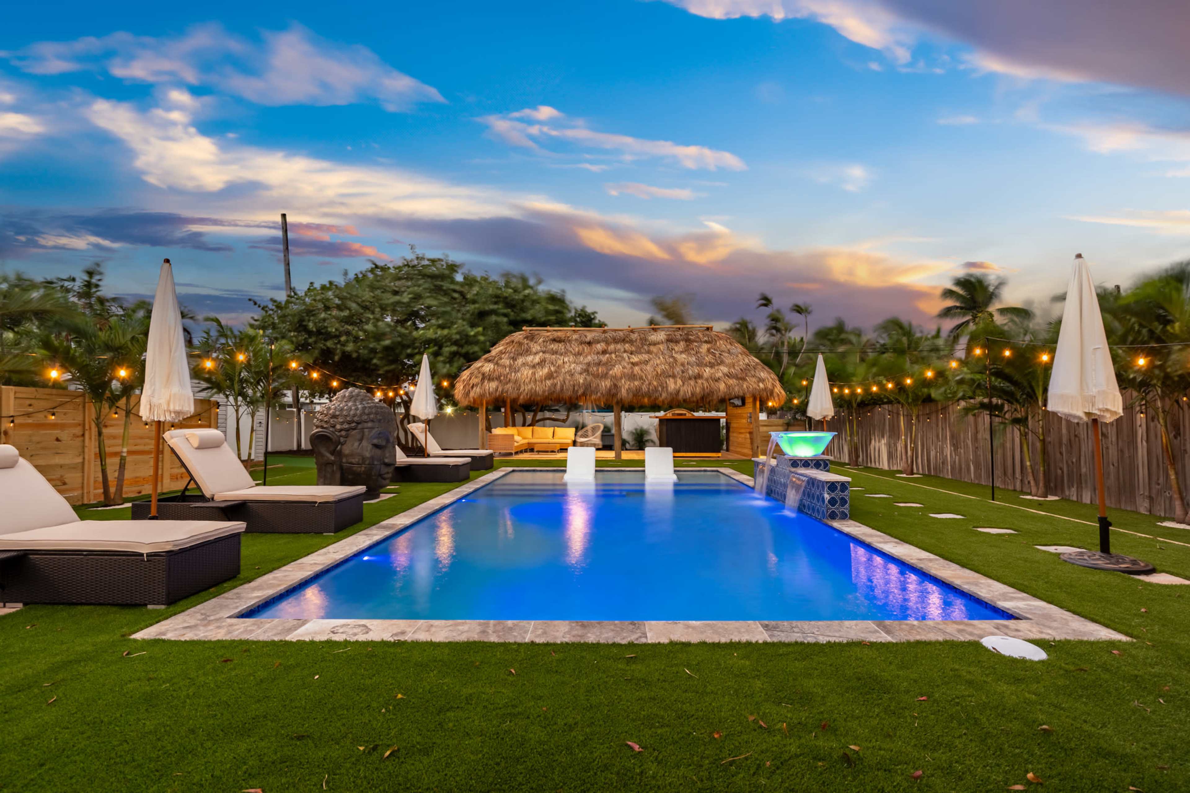 A swimming pool surrounded by lounge chairs and a thatched-roof gazebo is set in a green lawn under a colorful sky.
