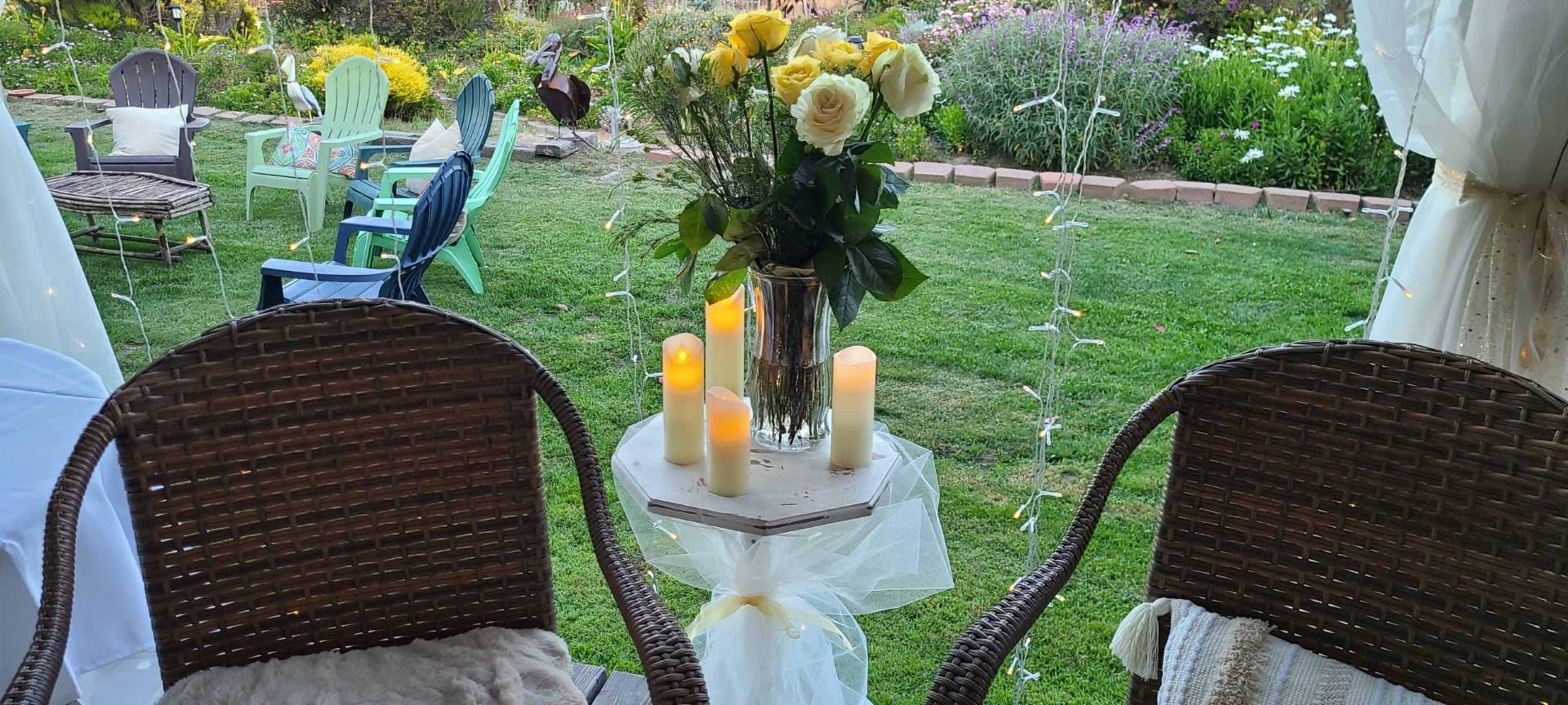 A table with a vase of yellow and white roses and candles stands between two wicker chairs in a garden setting.