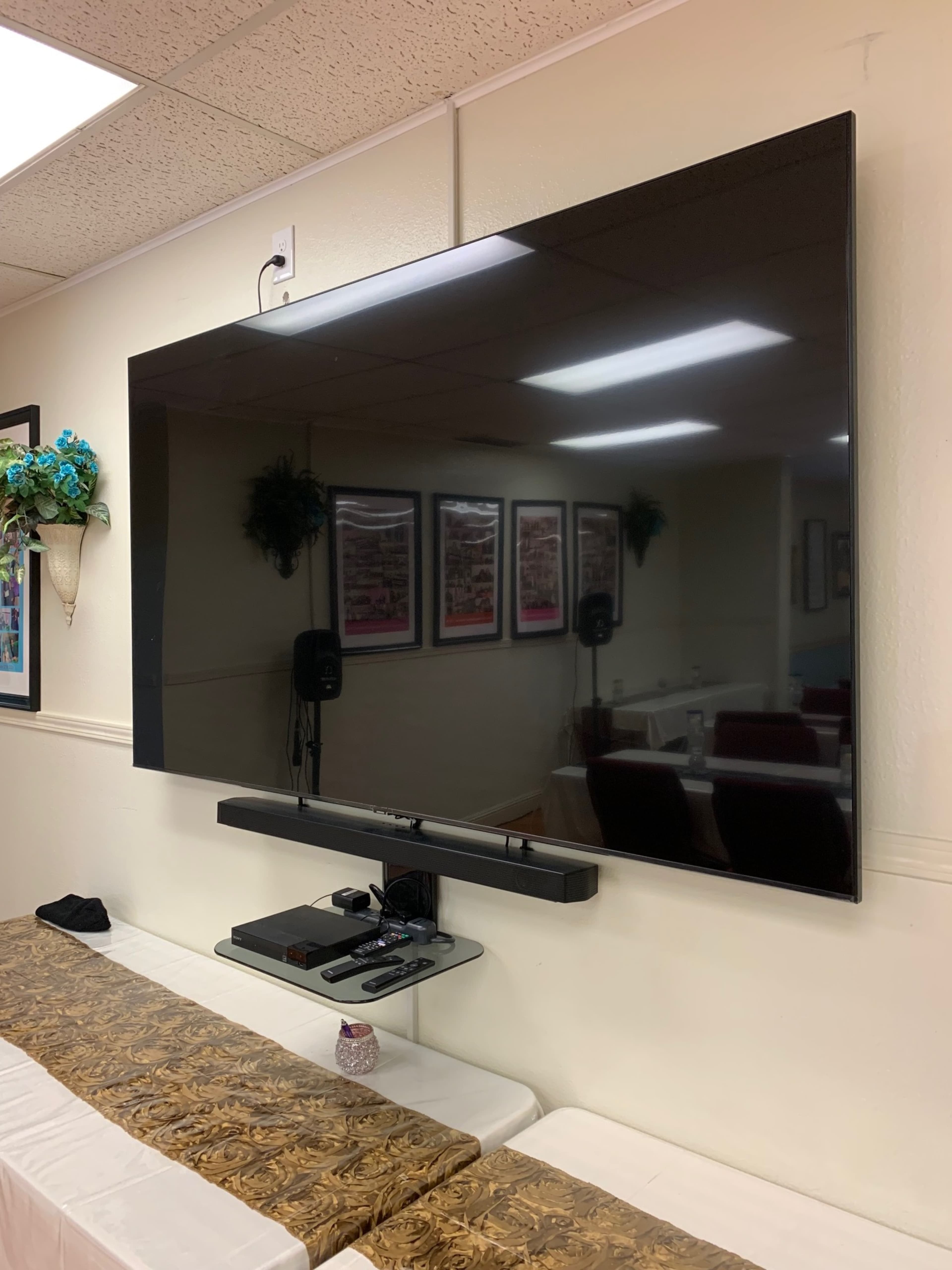A large flat-screen television is mounted on a wall above a low shelf with a soundbar and media devices, with seating and framed pictures visible in the background.