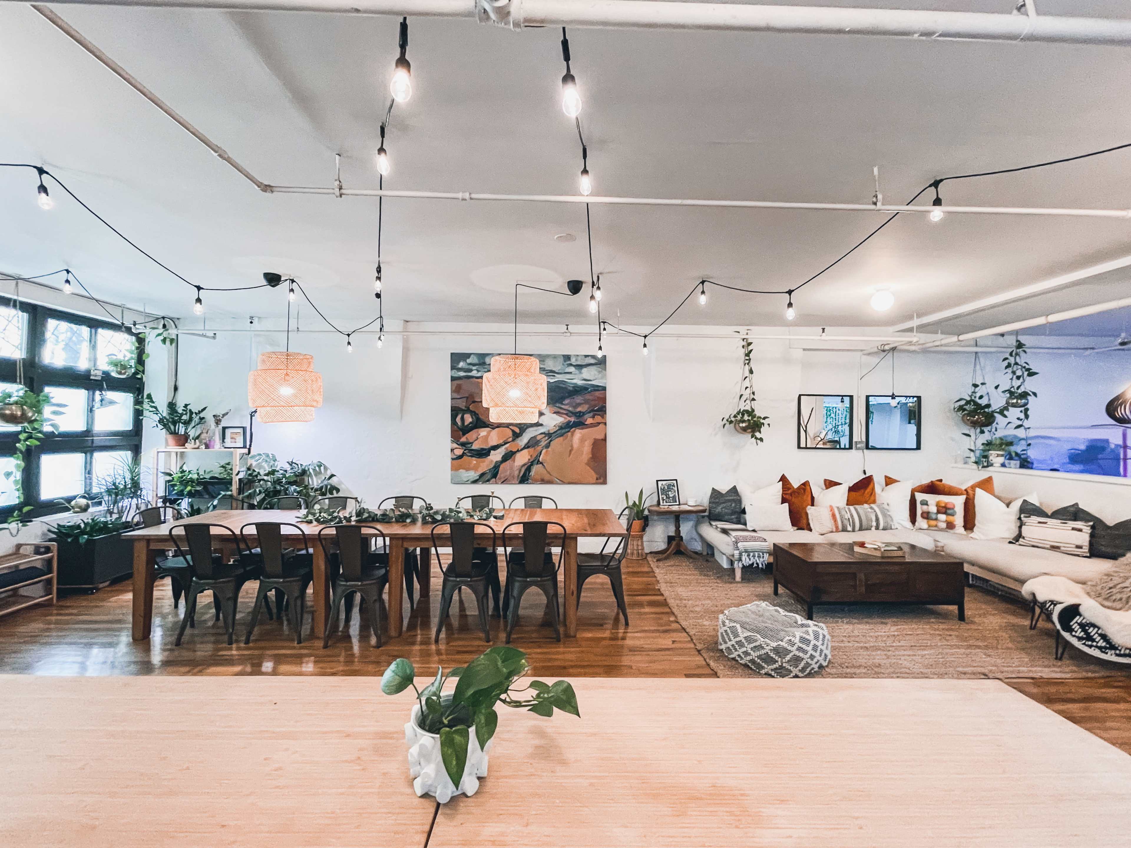 A spacious, light-filled living area with a large dining table, a cozy seating arrangement, decorative plants, and soft lighting fixtures.