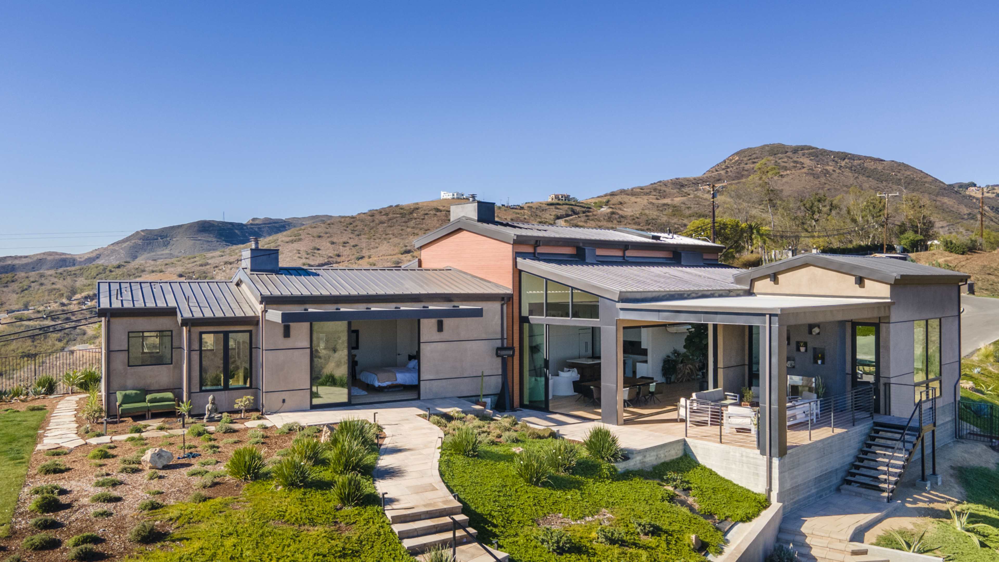 The image shows a modern house with large windows and a metal roof, situated on a hillside surrounded by landscaped greenery.
