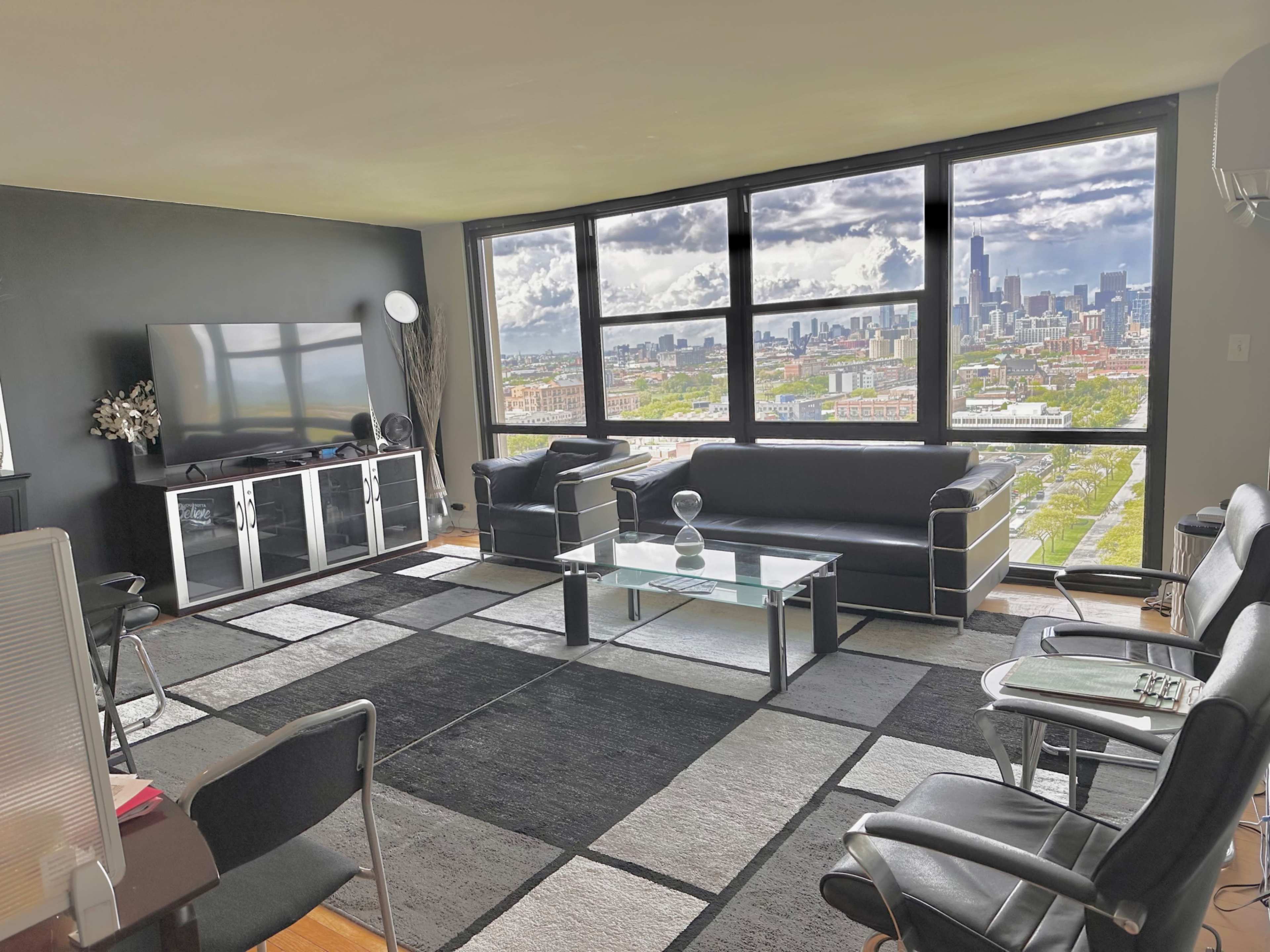 A modern living room features large windows offering a city skyline view, with black leather sofas, a glass coffee table, and a television on a cabinet.