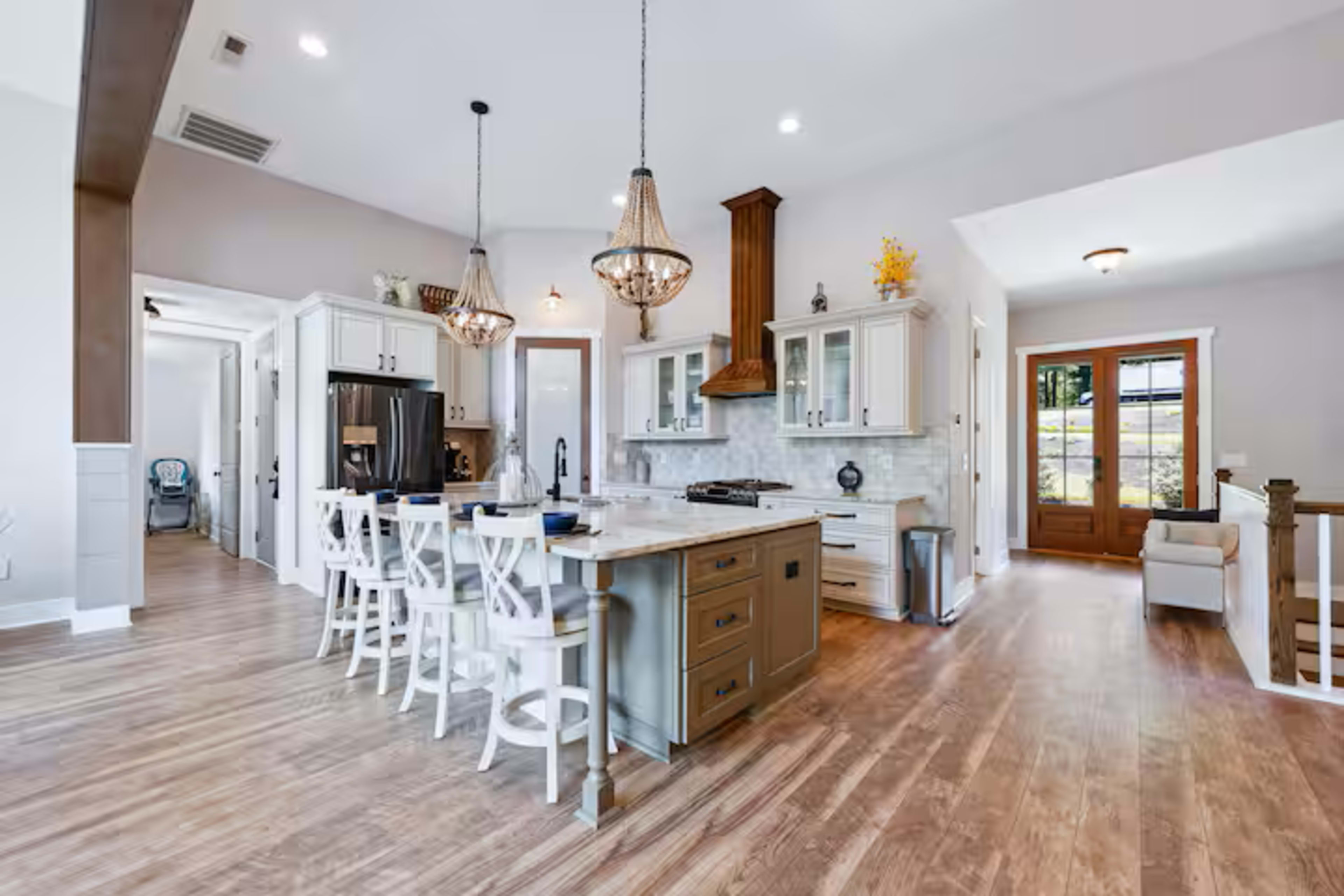The image shows a modern kitchen with a central island, white cabinetry, pendant lighting, and an adjoining living space.
