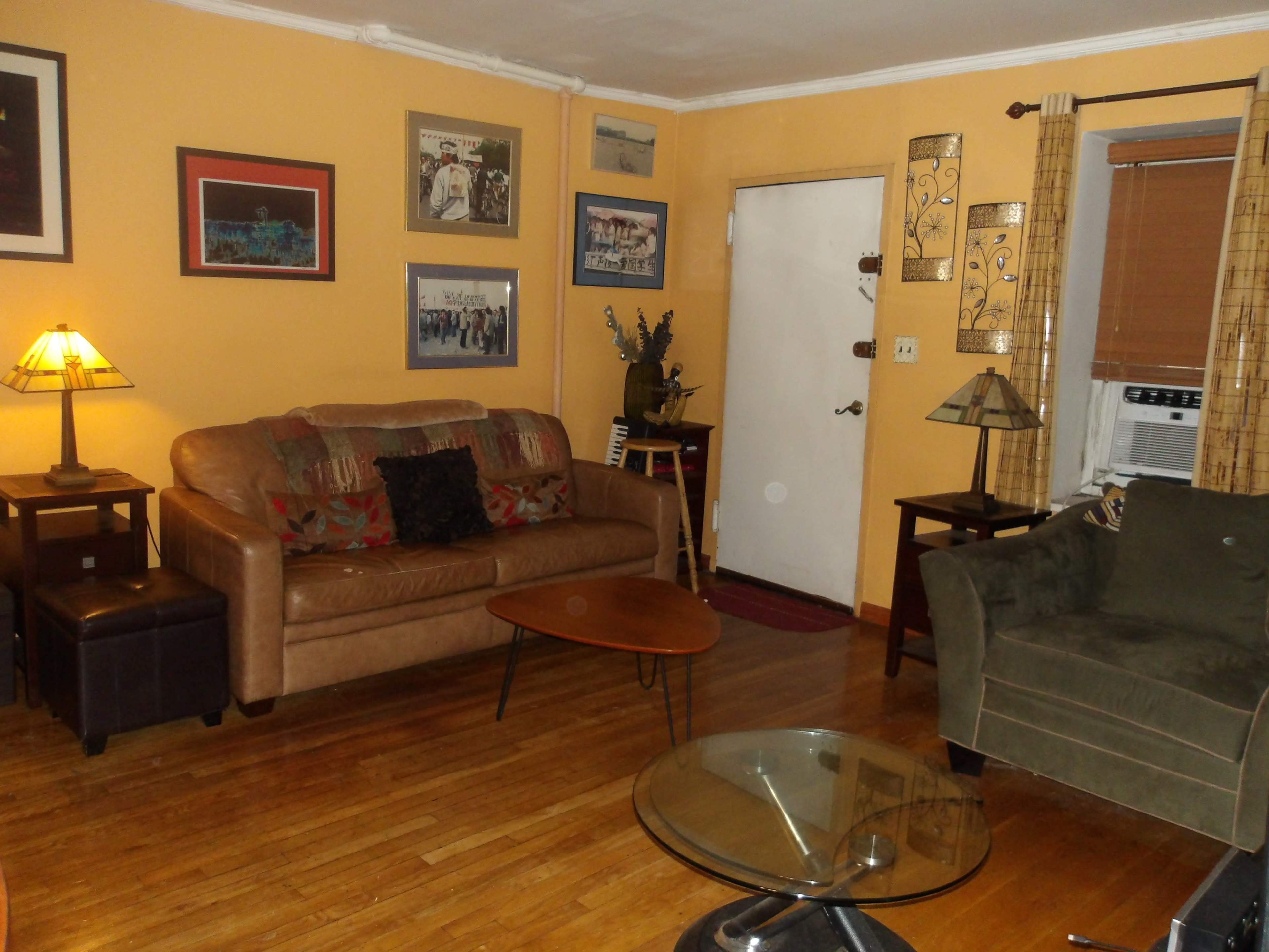 A living room with a brown leather sofa, green armchair, coffee table, and several framed pictures on the walls, illuminated by a lamp.