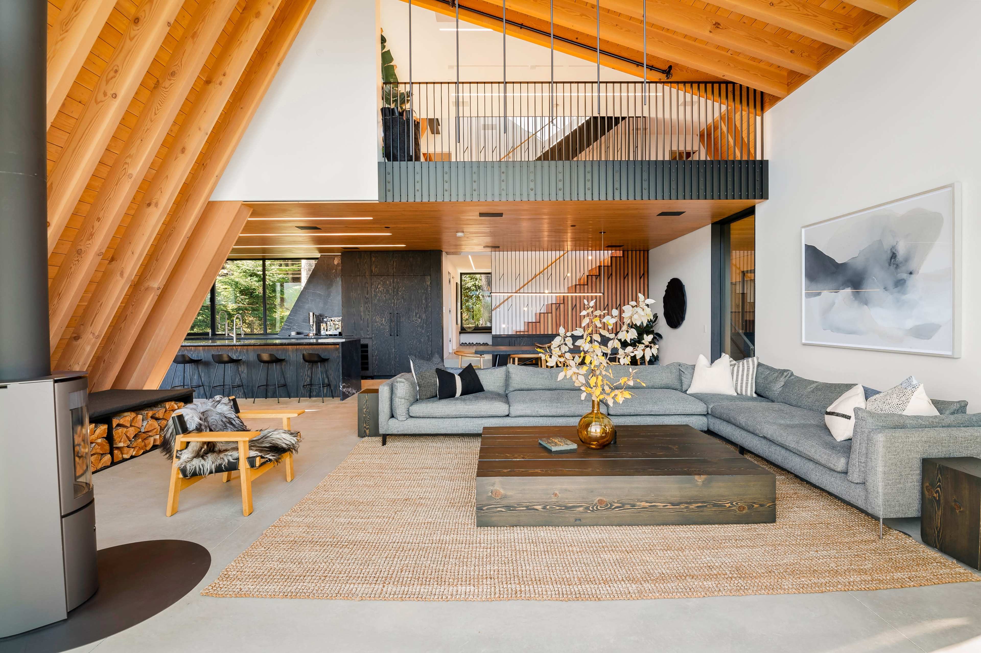 A modern living room with high wooden beams, a large gray sectional sofa, a central coffee table, and an open layout leading to a kitchen area.