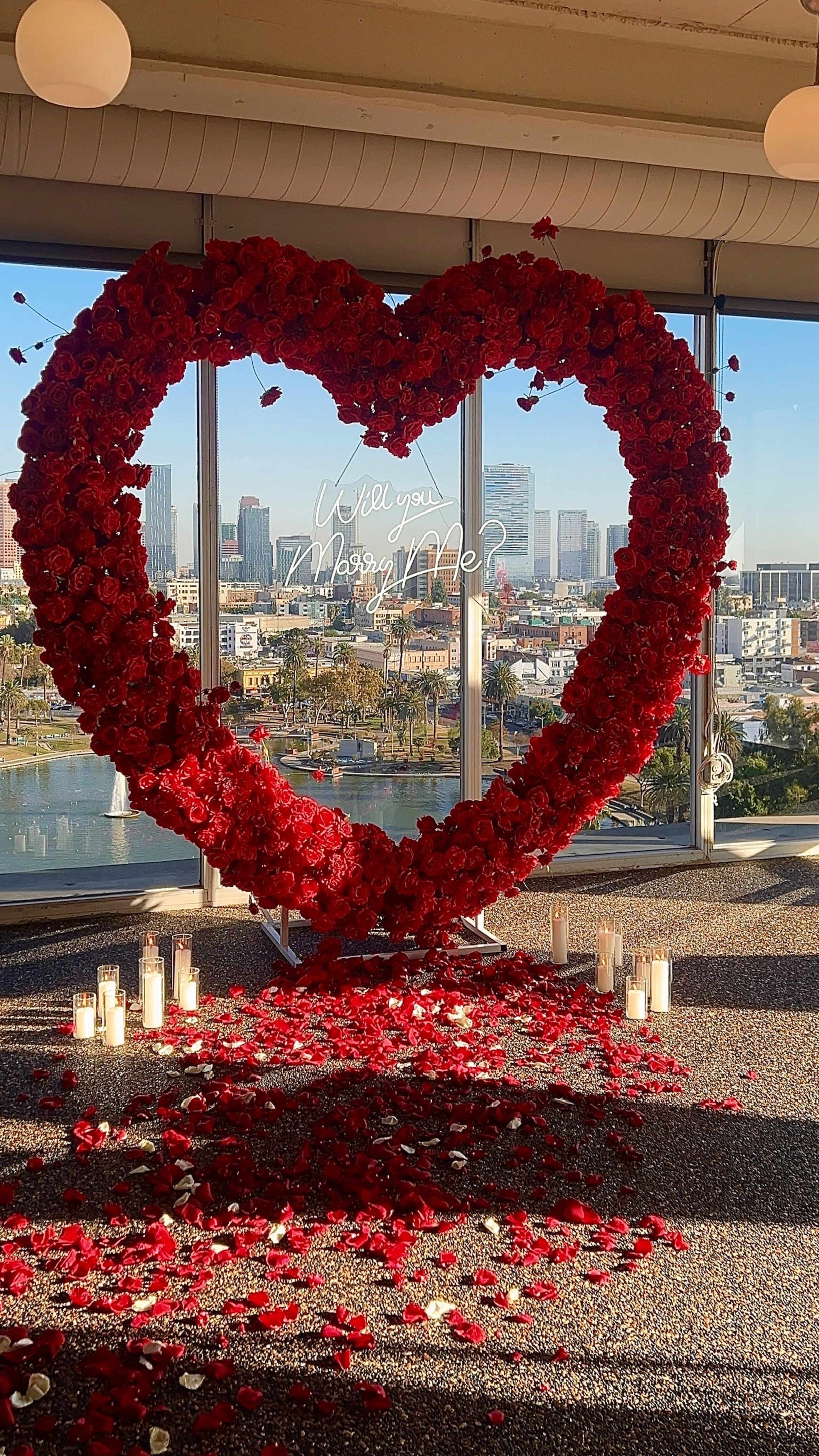 A large heart-shaped arrangement made of red roses is displayed in front of a panoramic city view, with scattered rose petals and candles on the floor.