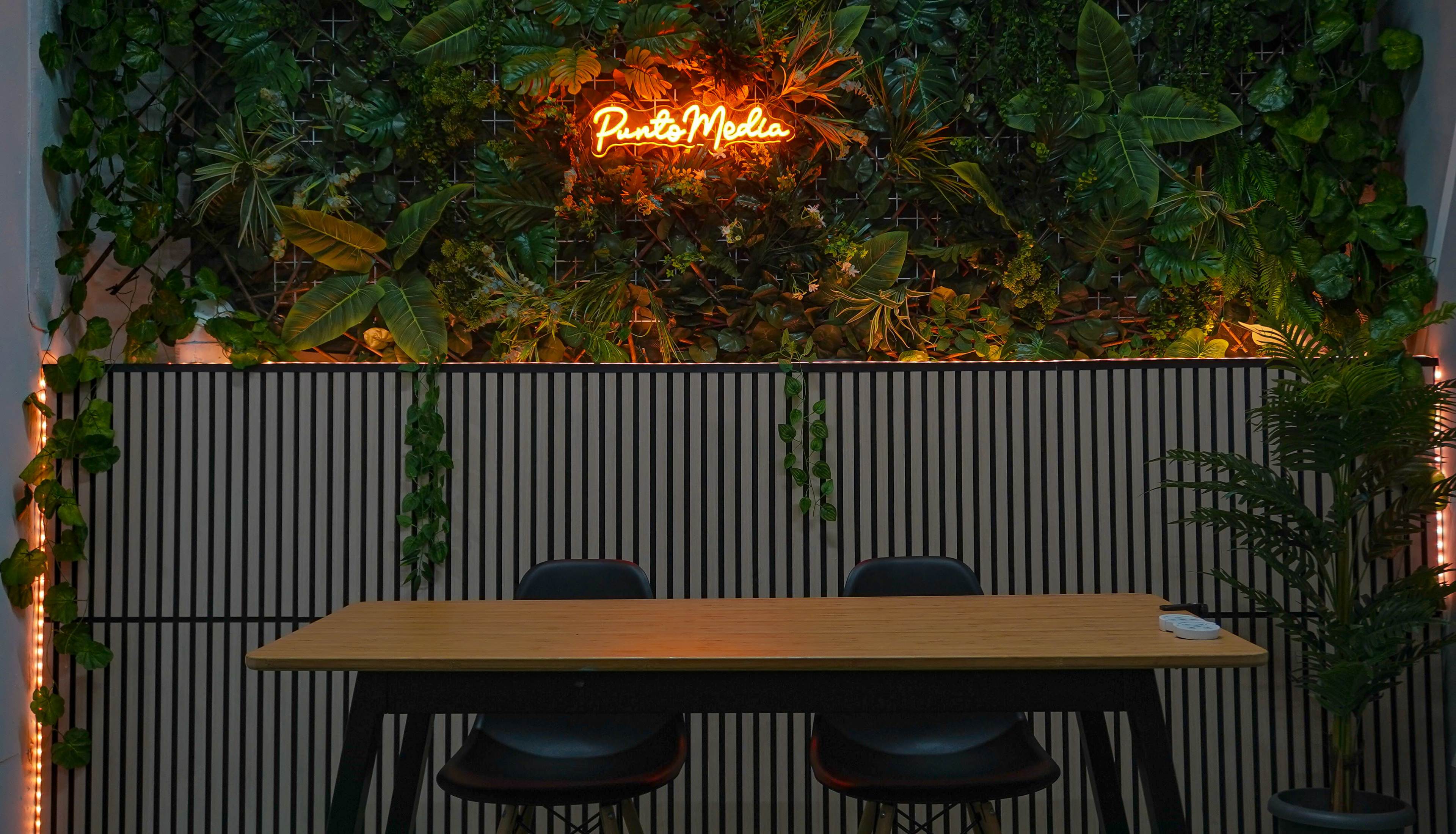 A wooden table with three black chairs is set against a lush green plant wall featuring an illuminated neon sign that reads "Punk Media."